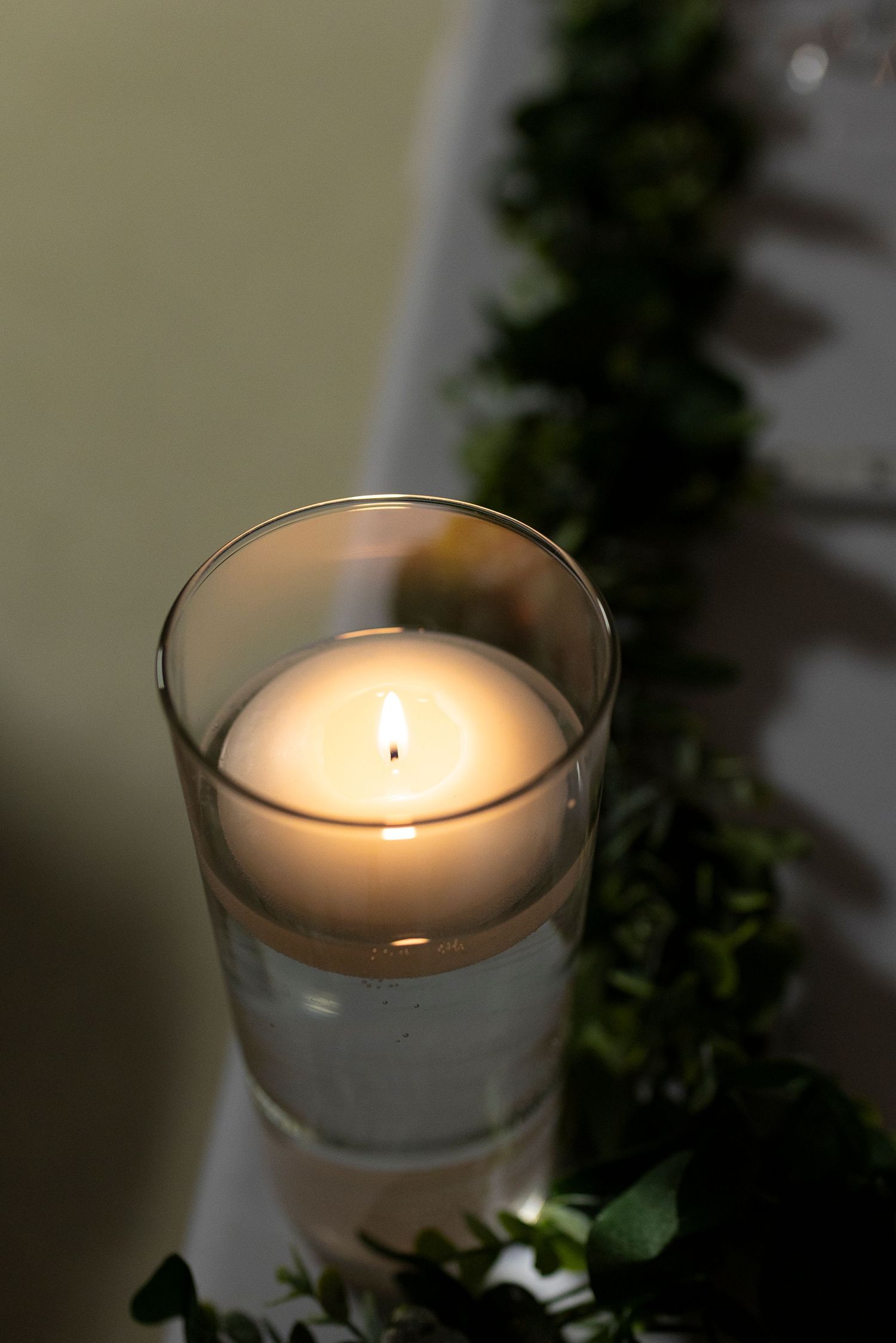 A lit floating candle in a glass vase surrounded by green garland decorations on a white table.