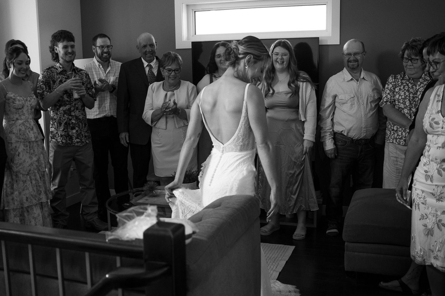 Black and white photo of a bride in backless gown signing wedding documents surrounded by smiling guests.