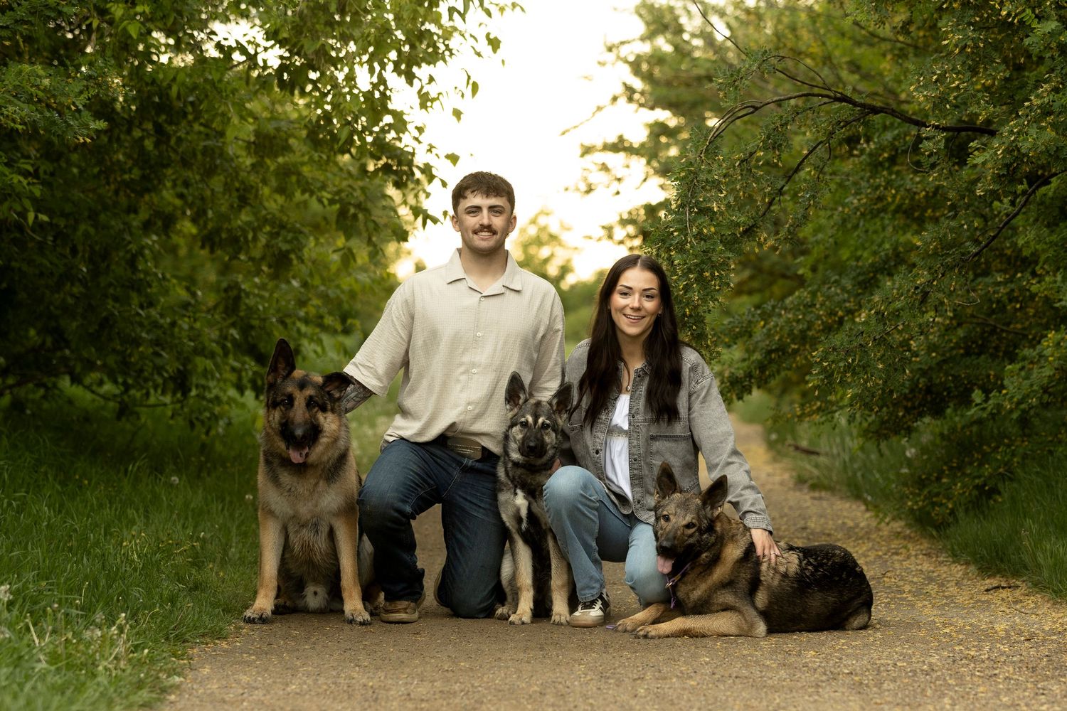 Couple kneeling on dirt path with three German Shepherds surrounded by lush green trees.
