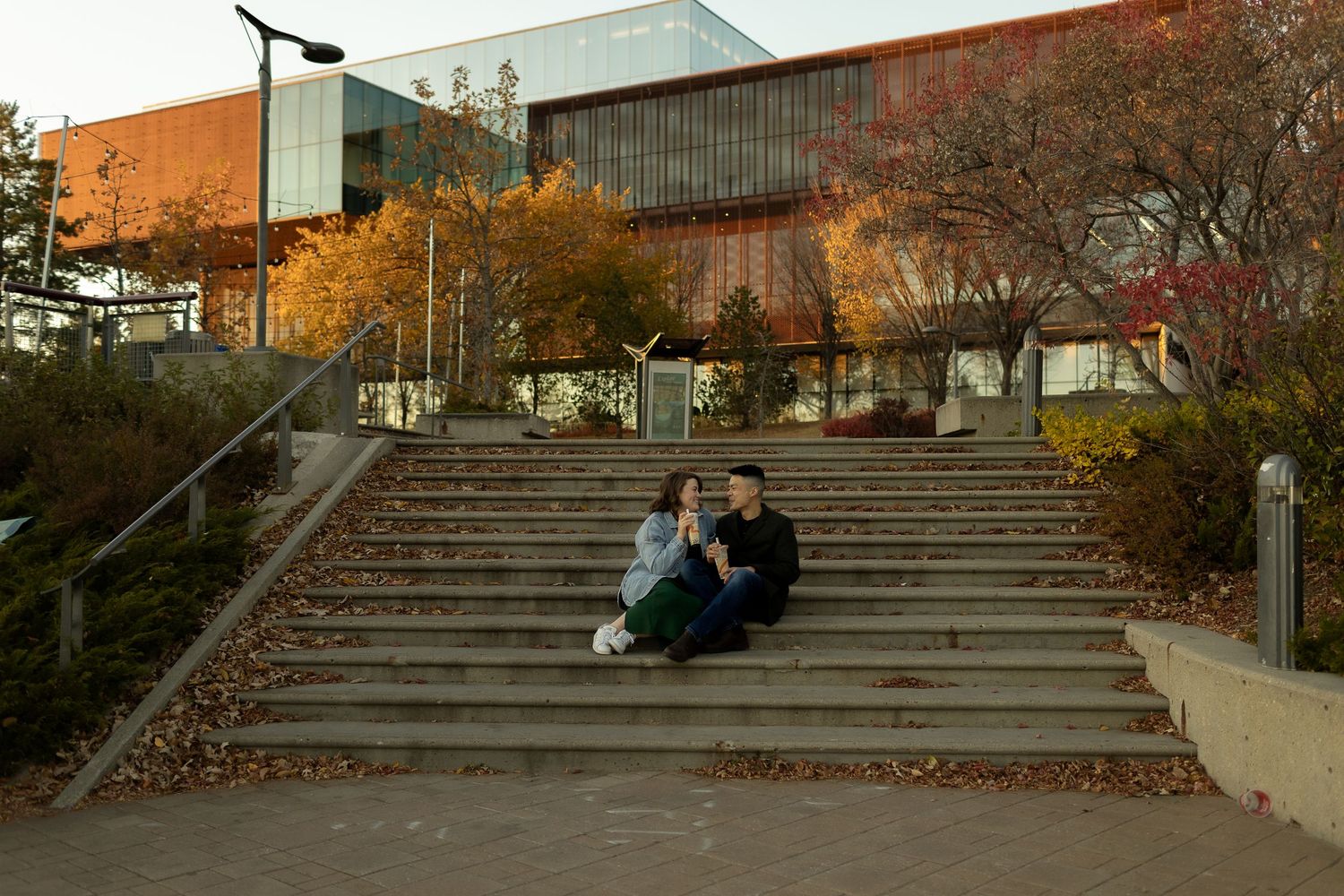 Couple sitting on outdoor steps near a modern building surrounded by autumn foliage.