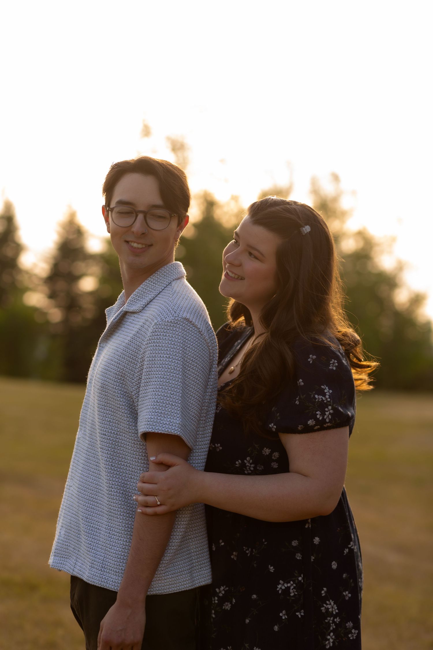 A couple embraces outdoors at golden hour, smiling warmly, with trees and soft sunlight in the background.