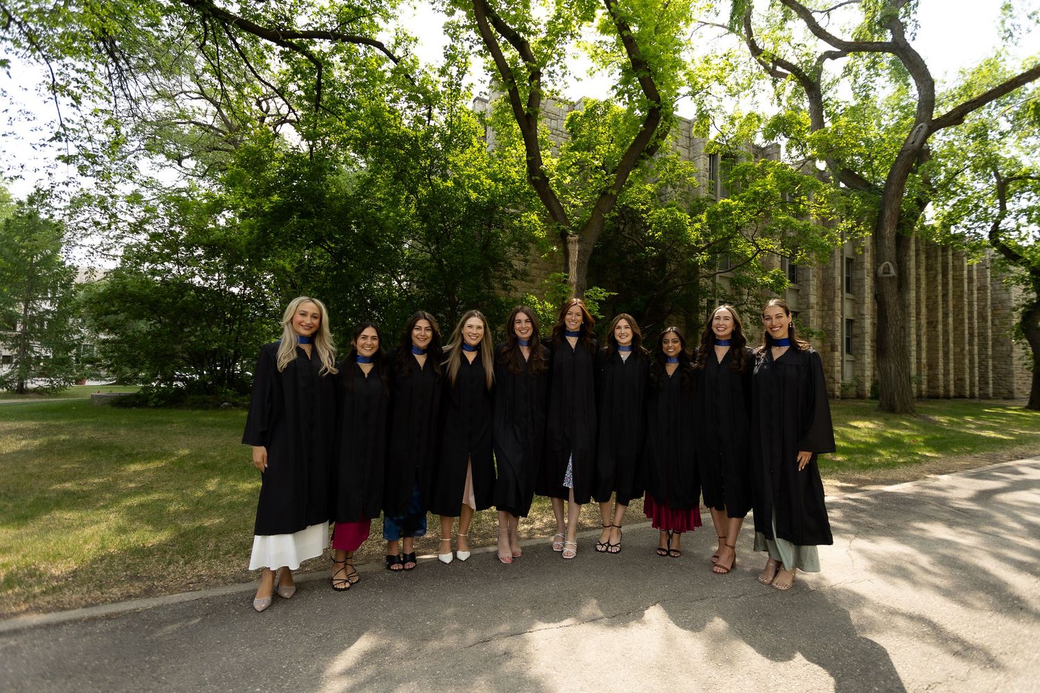 Group of graduates in black gowns posing outdoors on a sunny day near a historic building surrounded by trees.