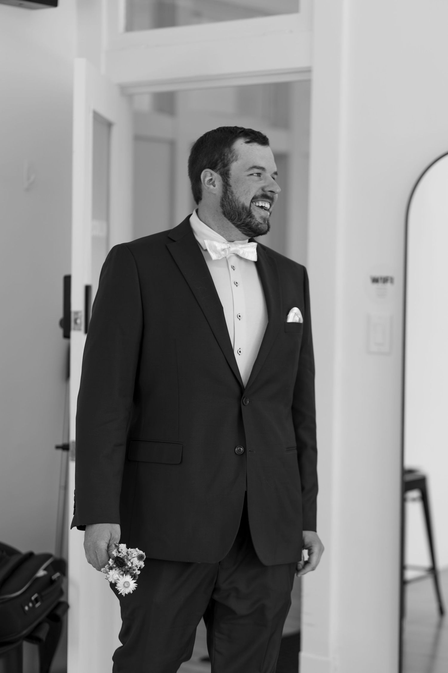 Smiling groom in black tuxedo with white bow tie and boutonniere, standing in a bright room on his wedding day.