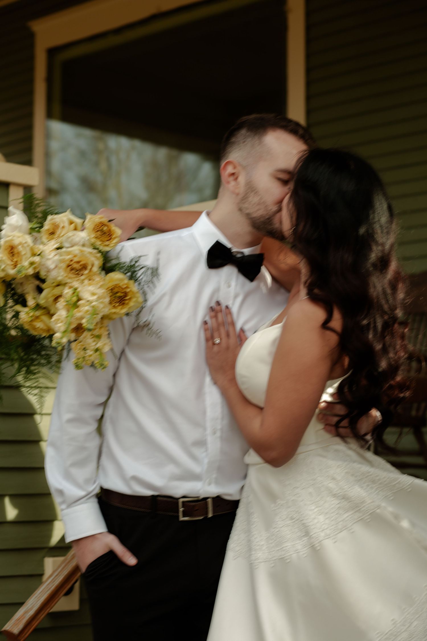 A bride and groom share a romantic kiss, she holds yellow roses bouquet, wearing white gown and he wears black bow tie.