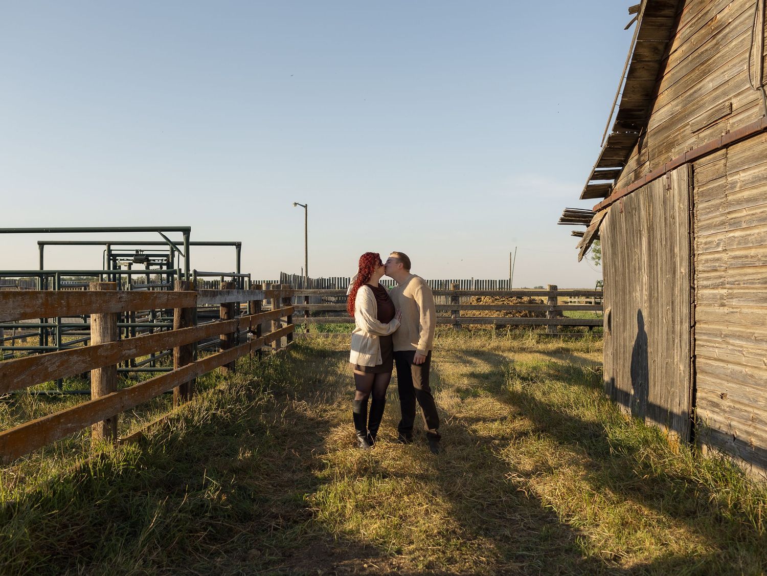 A couple embracing outside a rustic barn with wooden fences and open farmland at sunset in the Canadian prairies on a farm.