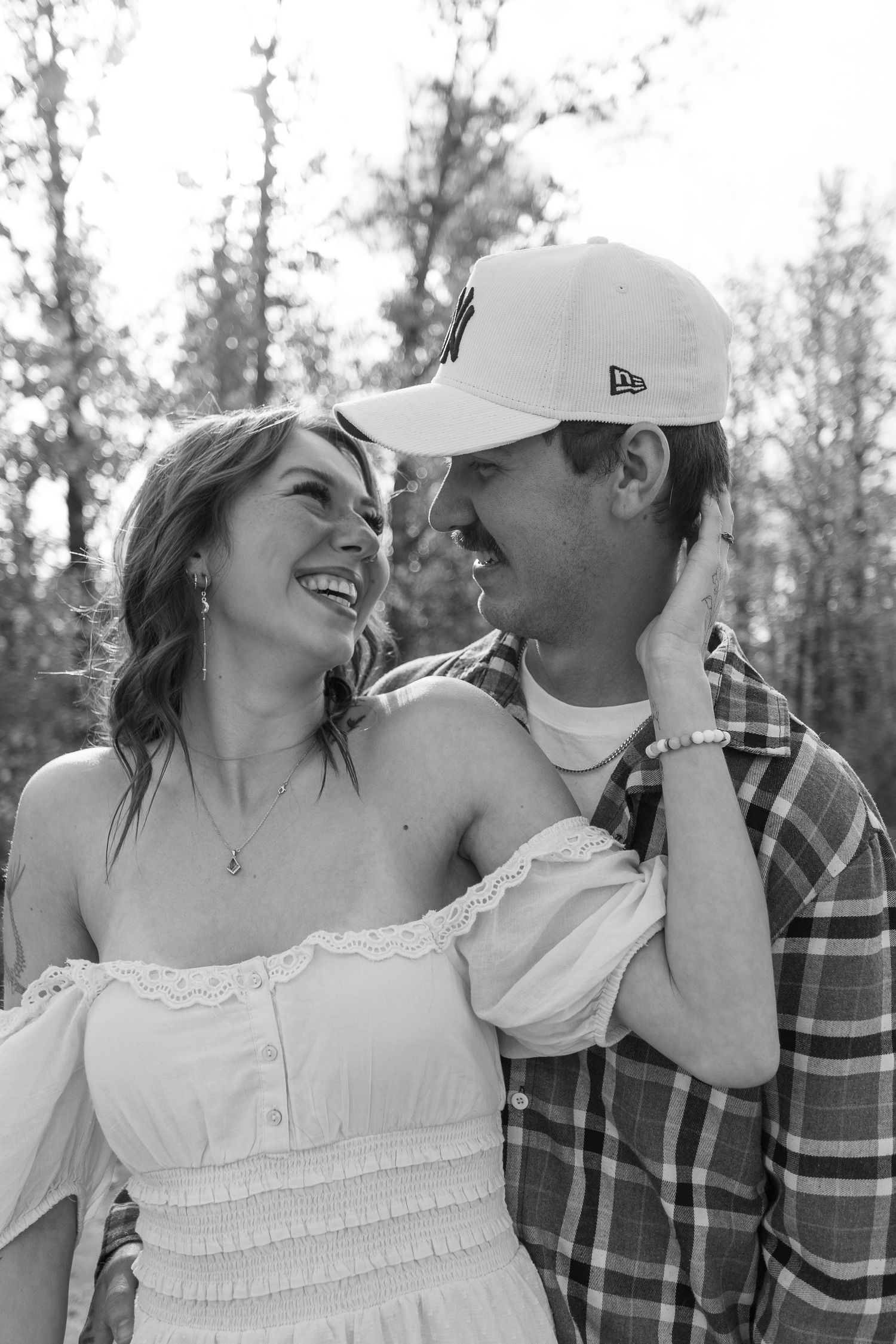 A romantic black and white portrait of a couple laughing together outdoors, with one wearing a baseball cap.