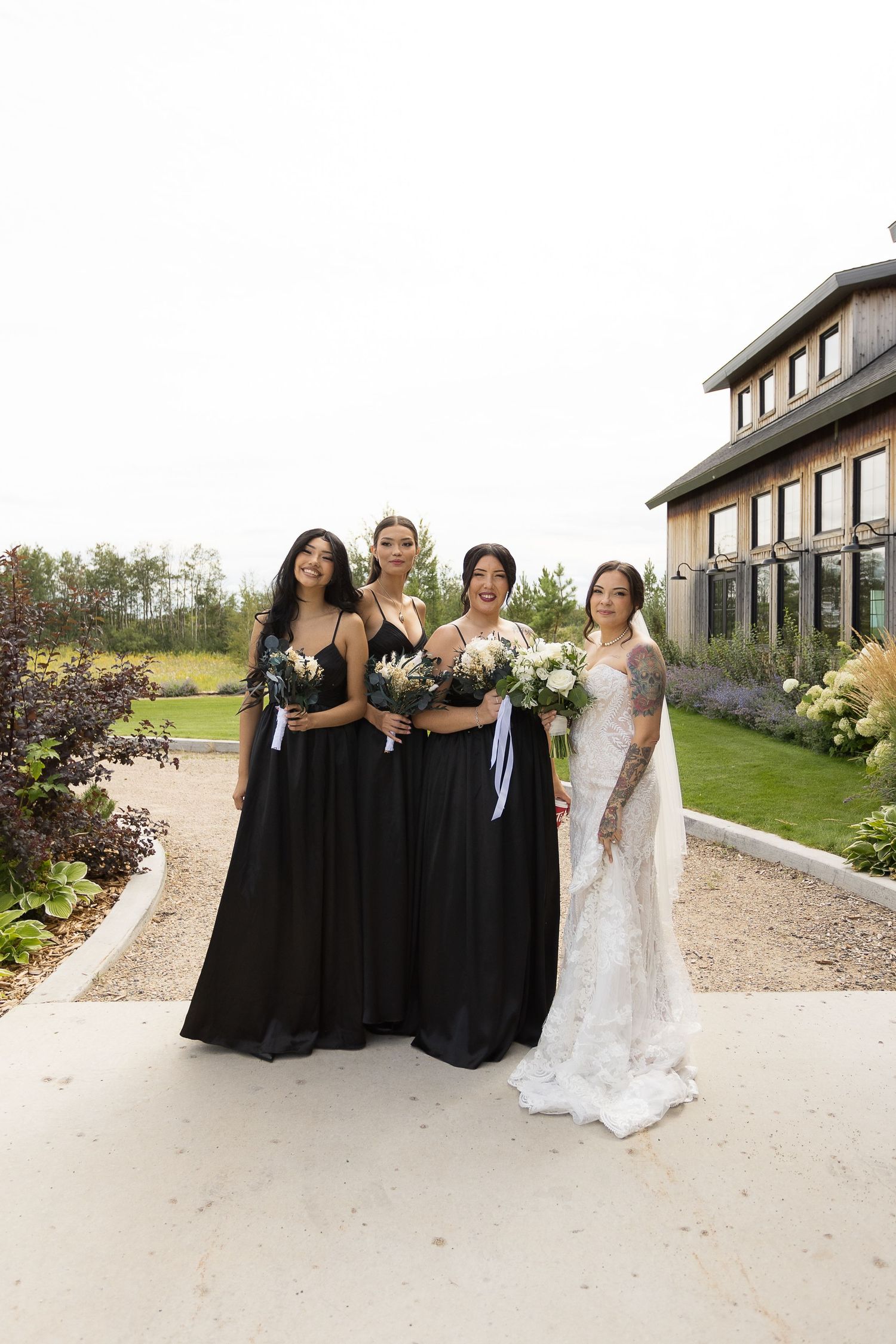 Wedding party in black and white dresses posing outside a modern venue on a gravel path at Barn at Wind's Edge in Saskatoon, Saskatchewan.