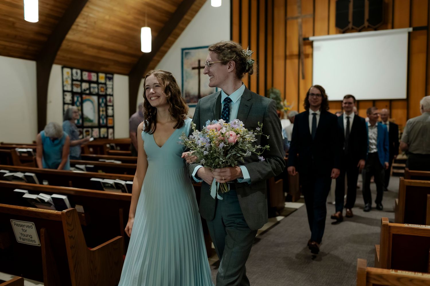Wedding party members walking down the aisle in a traditional church setting with wood paneled walls.