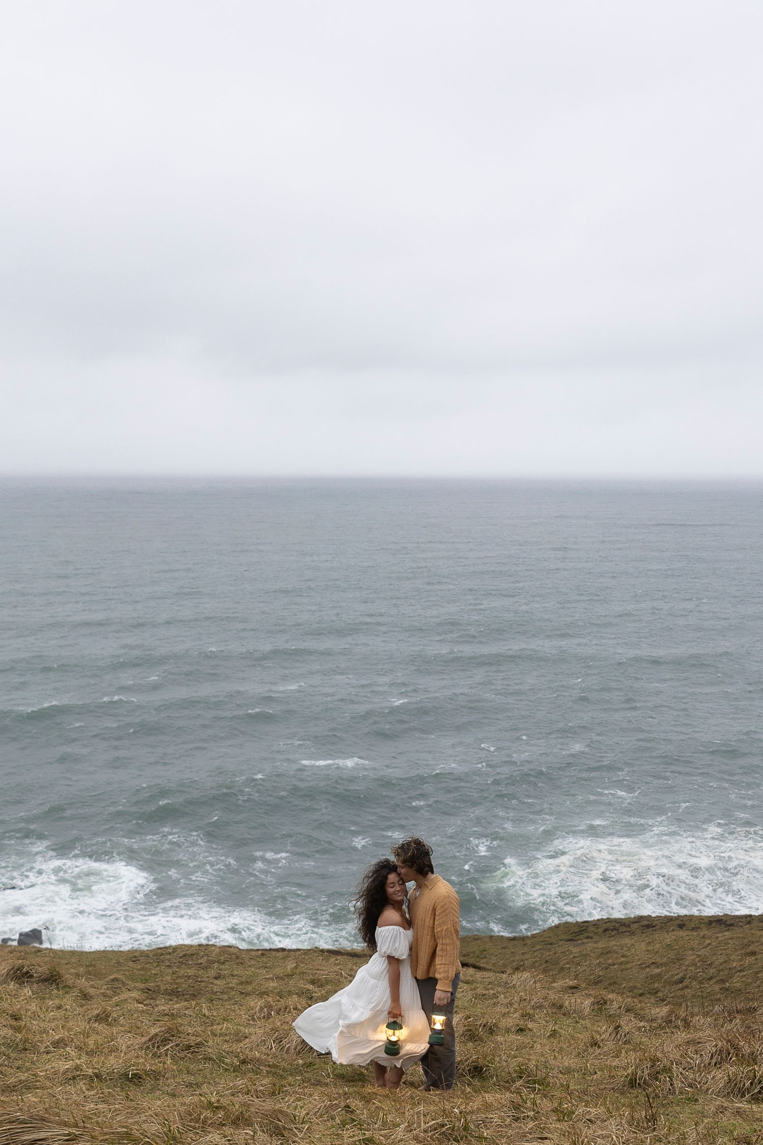 A couple embraces on a dramatic coastal cliff overlooking stormy ocean waters.