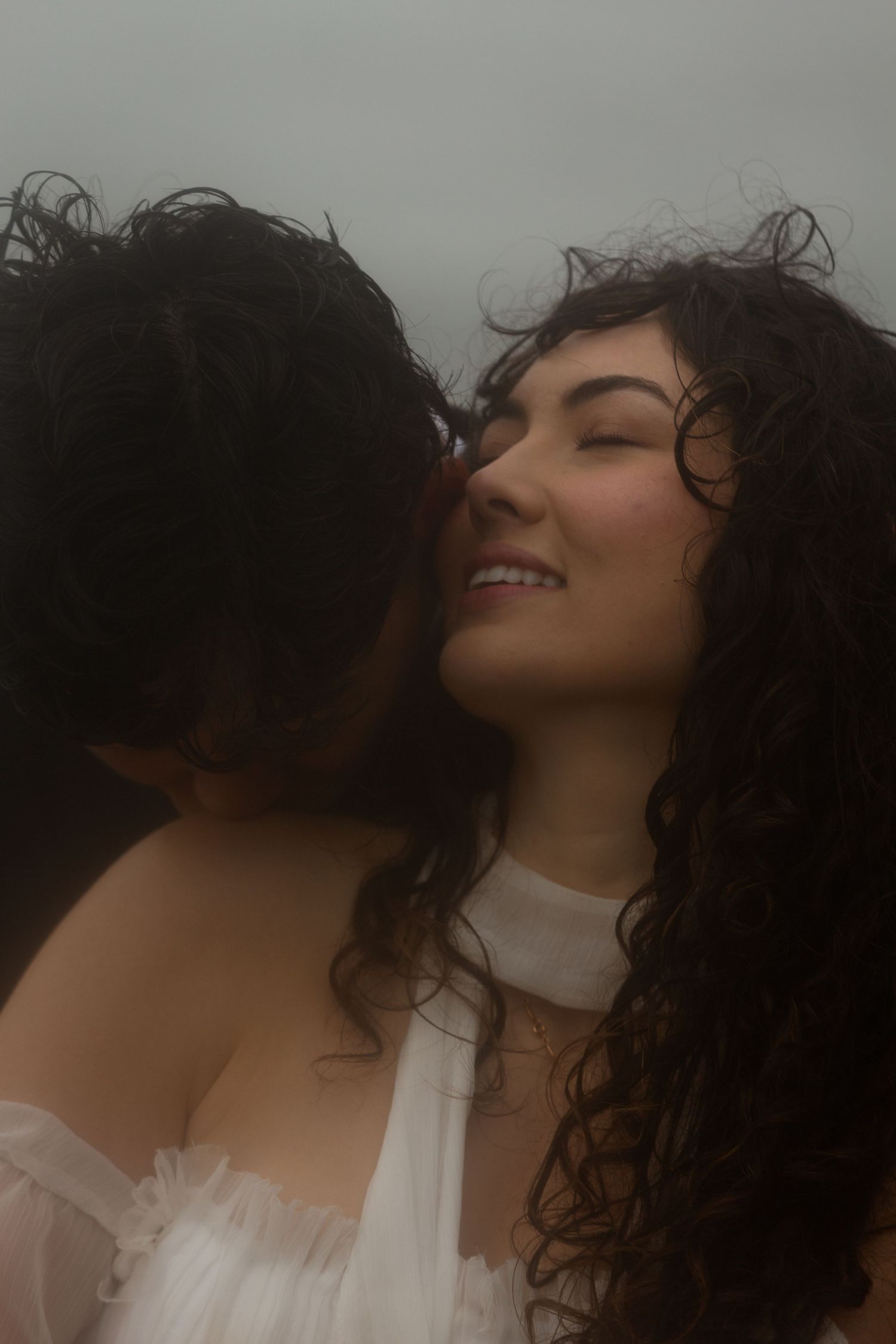 A smiling woman with curly dark hair in a white dress receives a kiss on the cheek in a romantic outdoor portrait.
