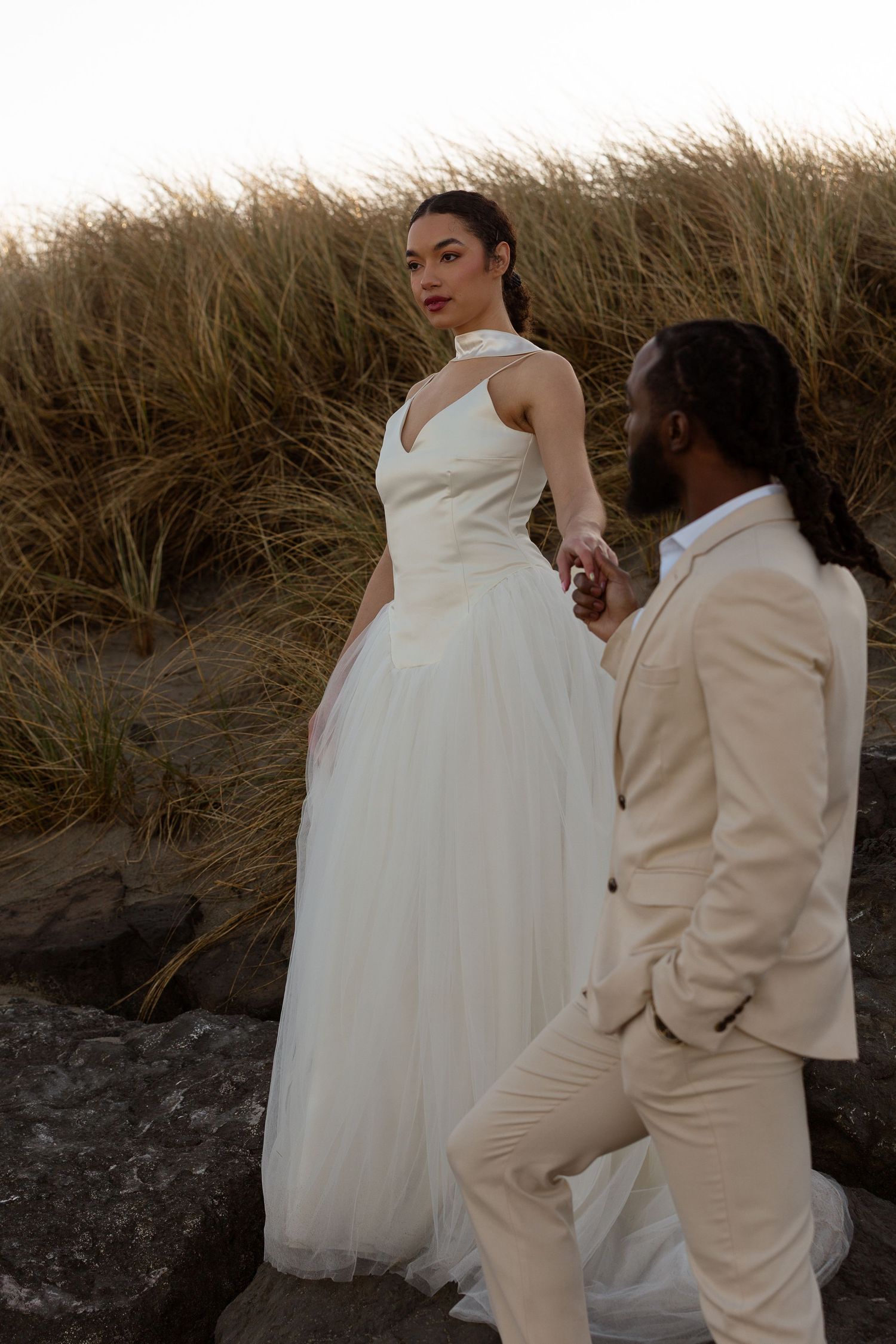 Interracial couple in wedding attire holding hands on rocky beach, bride in white gown, groom in beige suit.