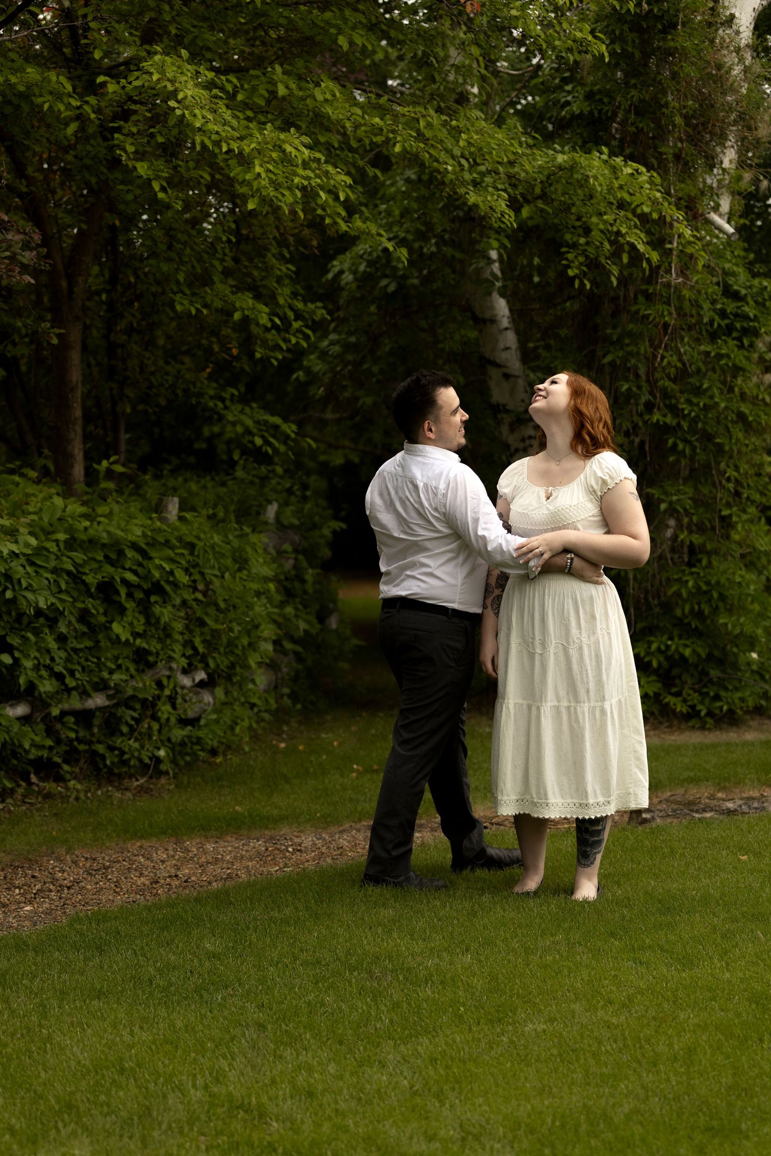 A couple laughs and dances together on a green lawn surrounded by lush garden trees.