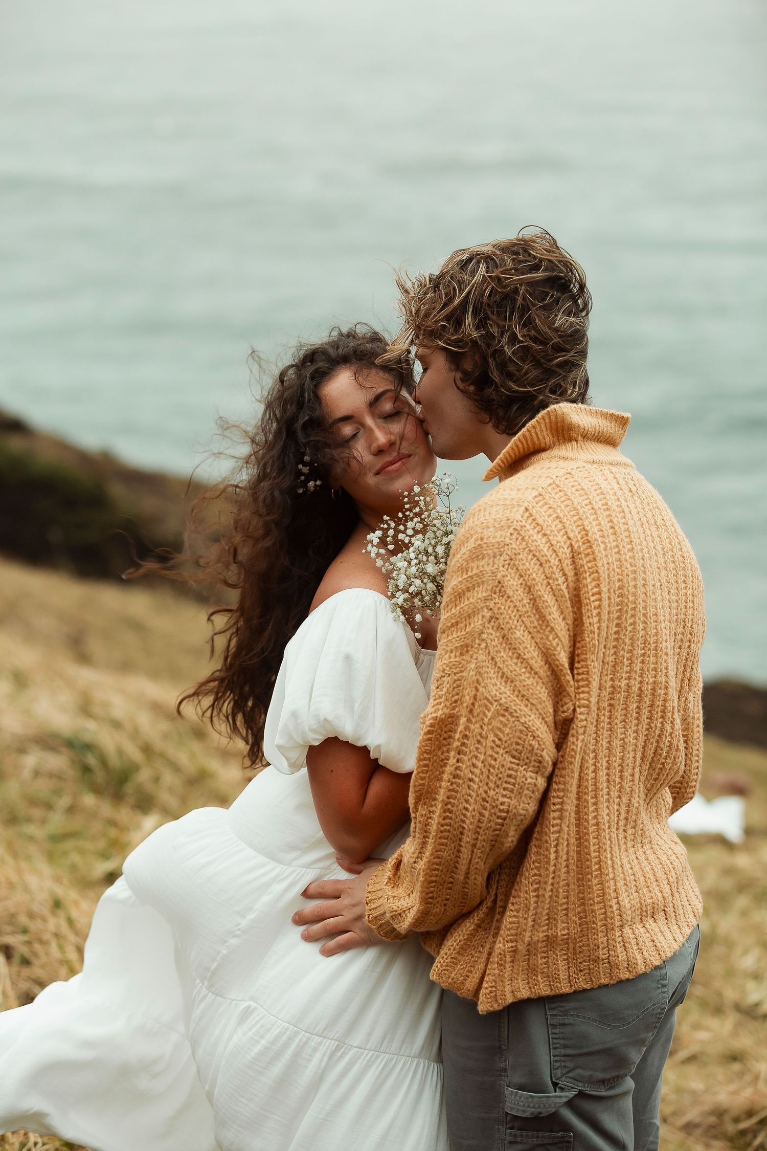 A couple shares intimate moments on a coastal cliff wearing casual knit sweaters.