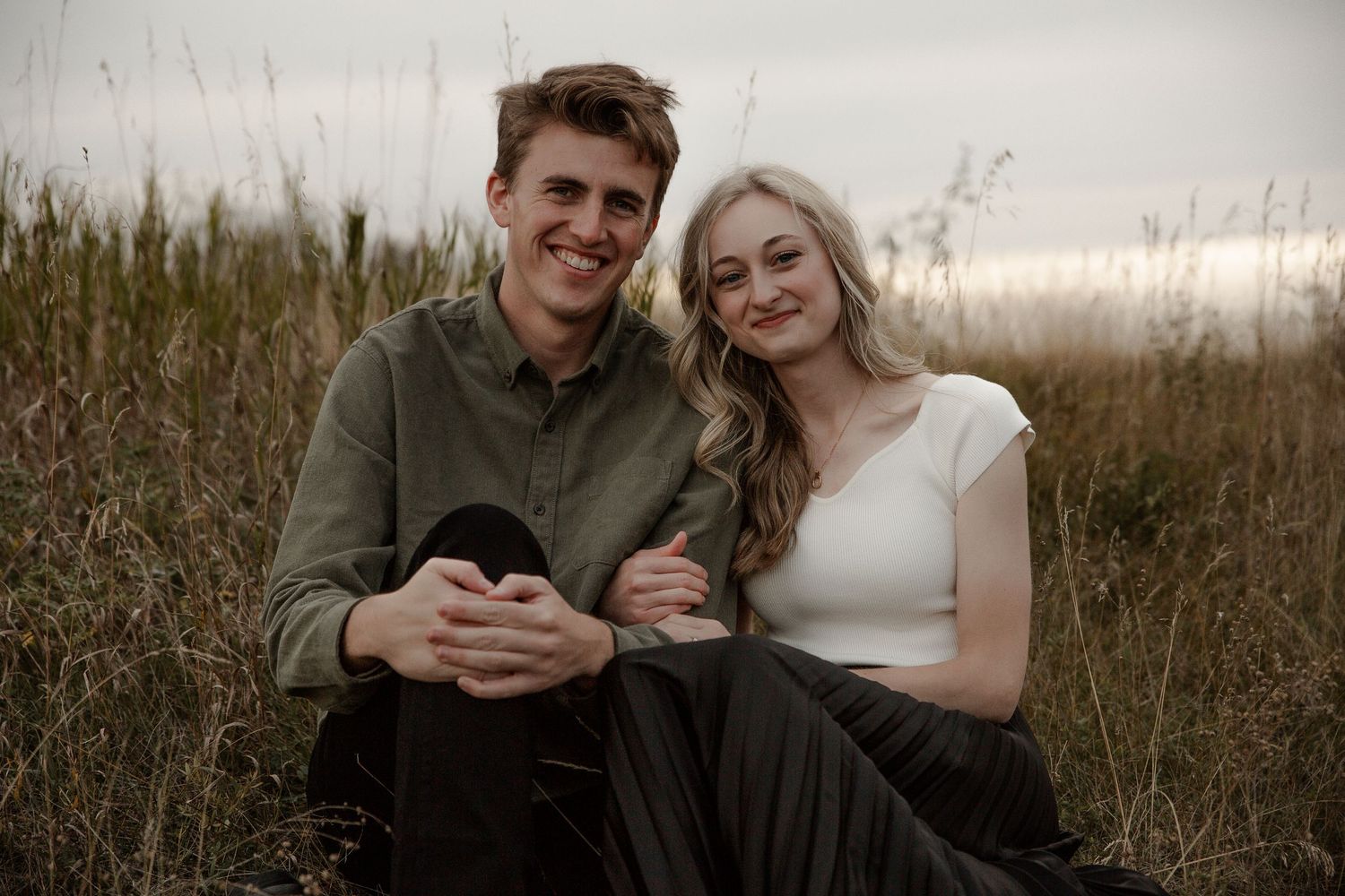 A young couple sits together smiling in a golden grass field, the woman in white top and man in olive shirt.