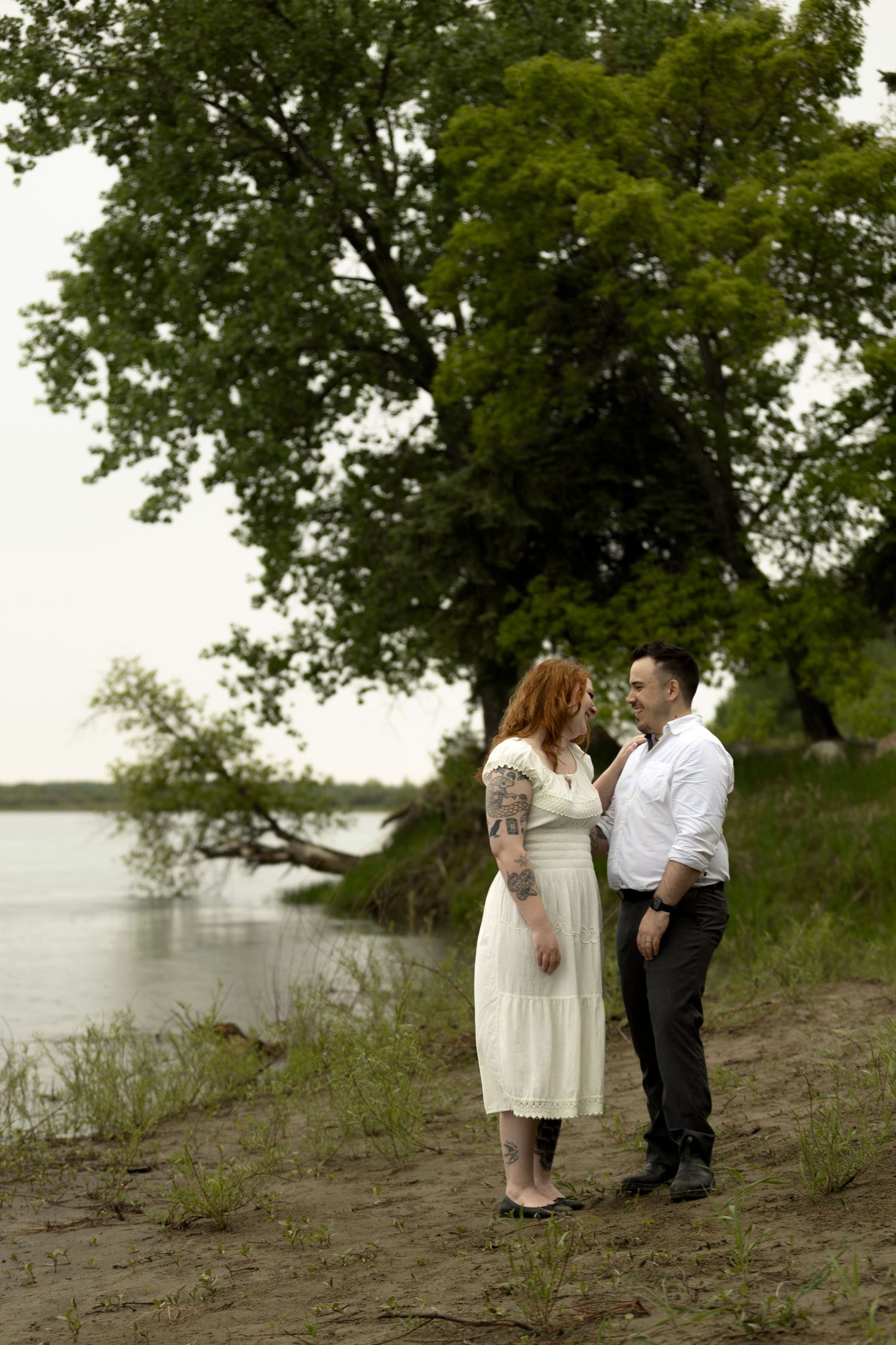 A tattooed woman in white dress and man in white shirt share a tender moment by a riverside.