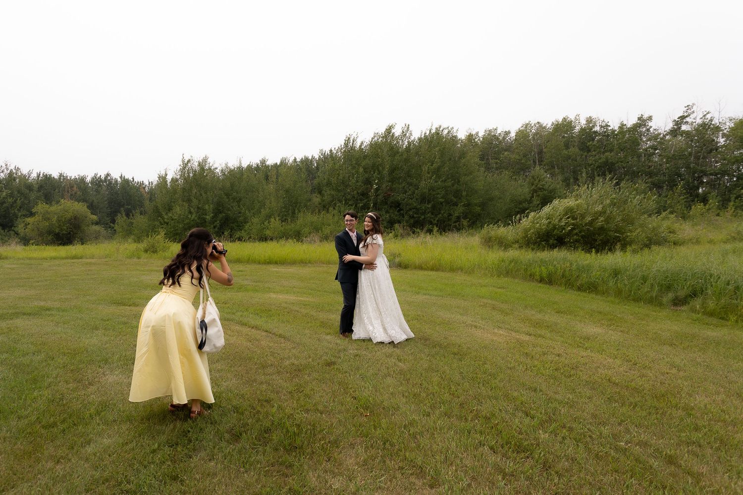 A photographer captures a wedding couple in a grassy field while wearing a yellow dress.