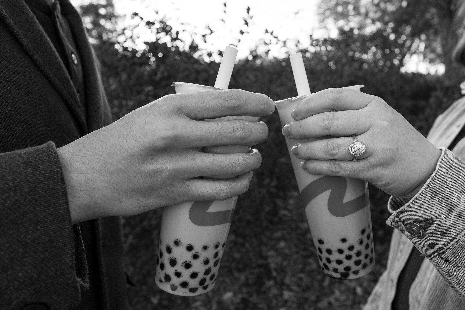 Two people toasting with boba tea drinks outdoors in a black and white photo, one wearing a ring.