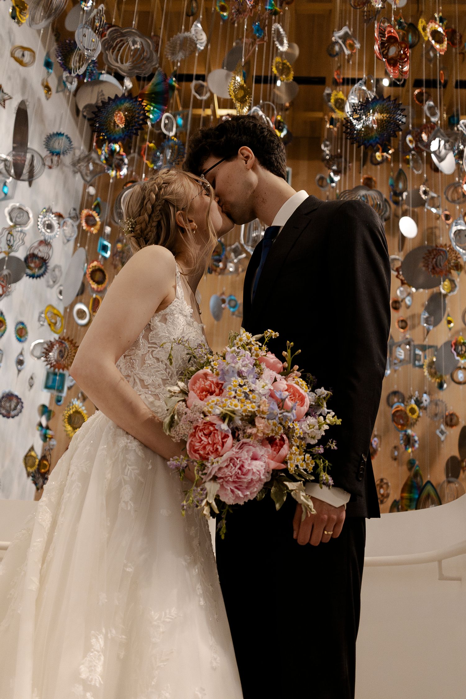 Bride and groom sharing a romantic kiss in wedding attire, with a colorful hanging art installation backdrop.