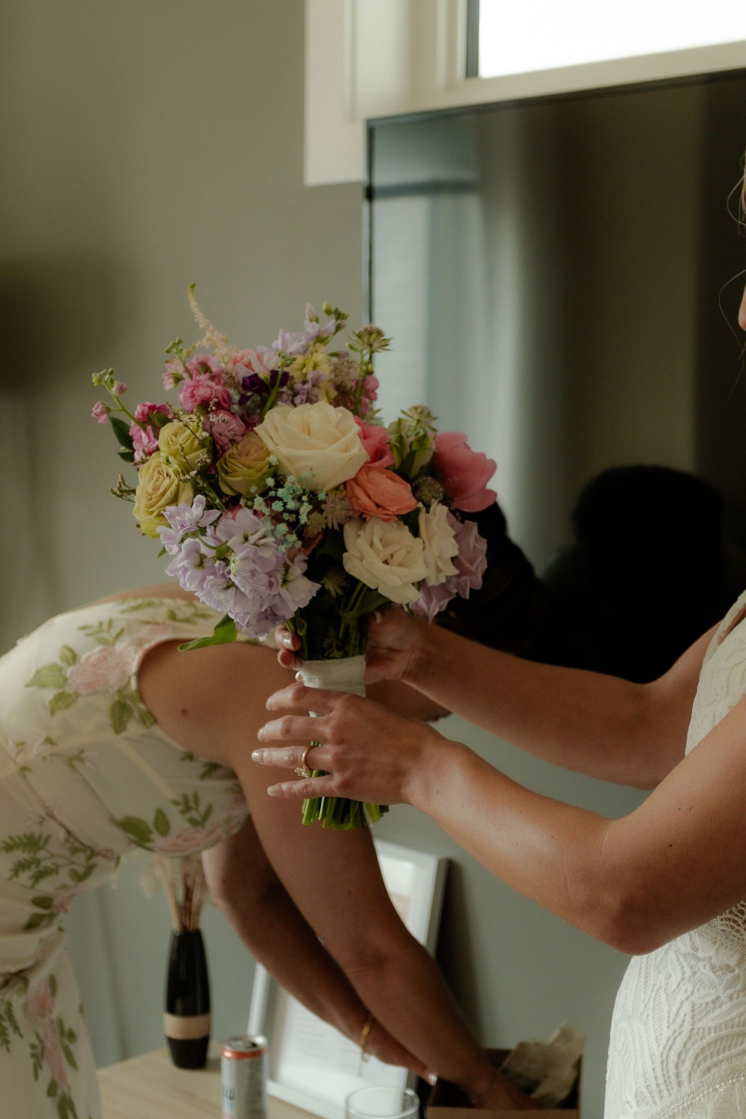 Bride in lace gown receives colorful wedding bouquet with pink, lavender, and cream flowers from bridesmaid near mirror.