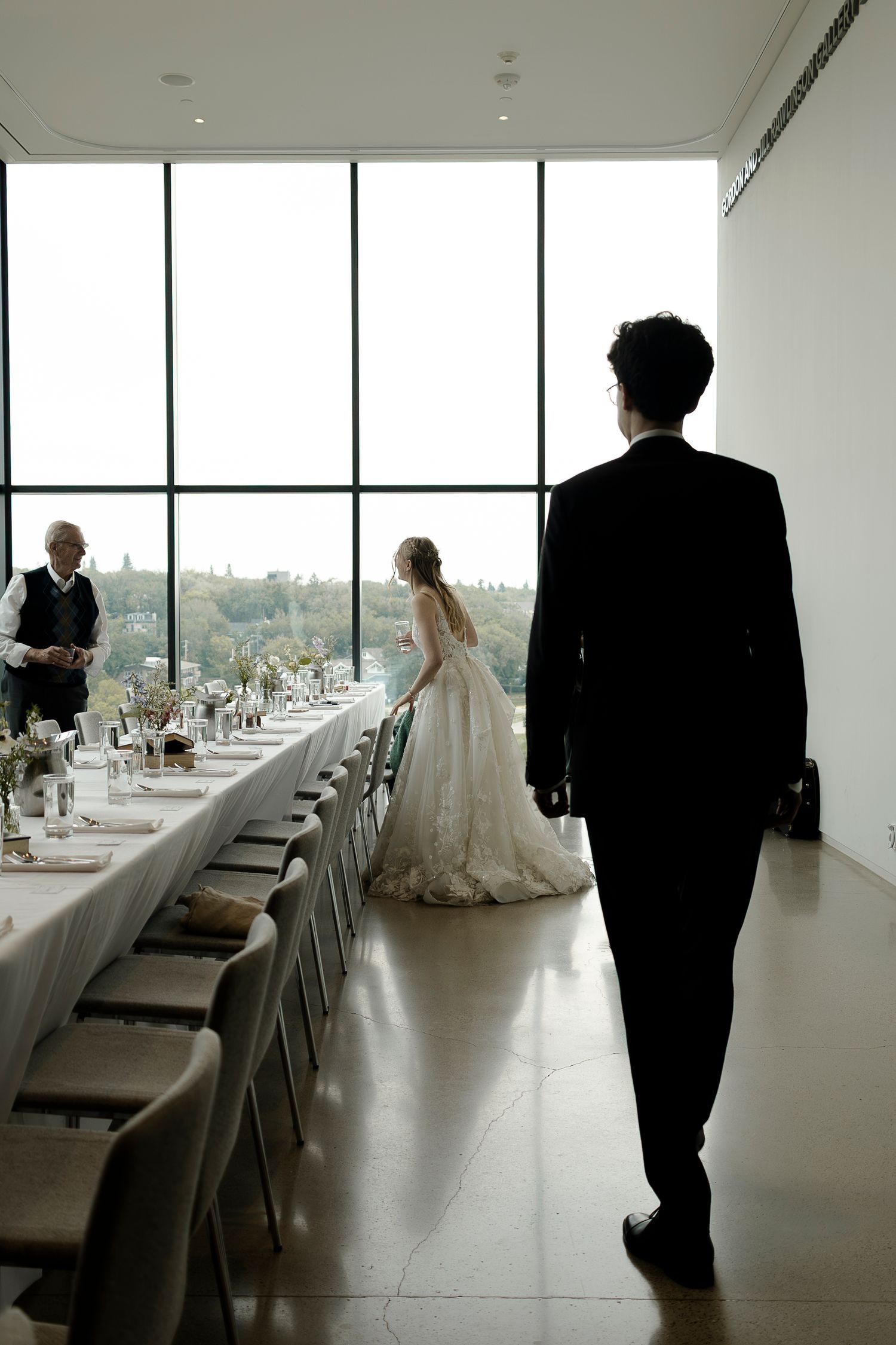 Groom in black suit walks toward bride in white gown standing by floor-to-ceiling windows in elegant wedding venue.