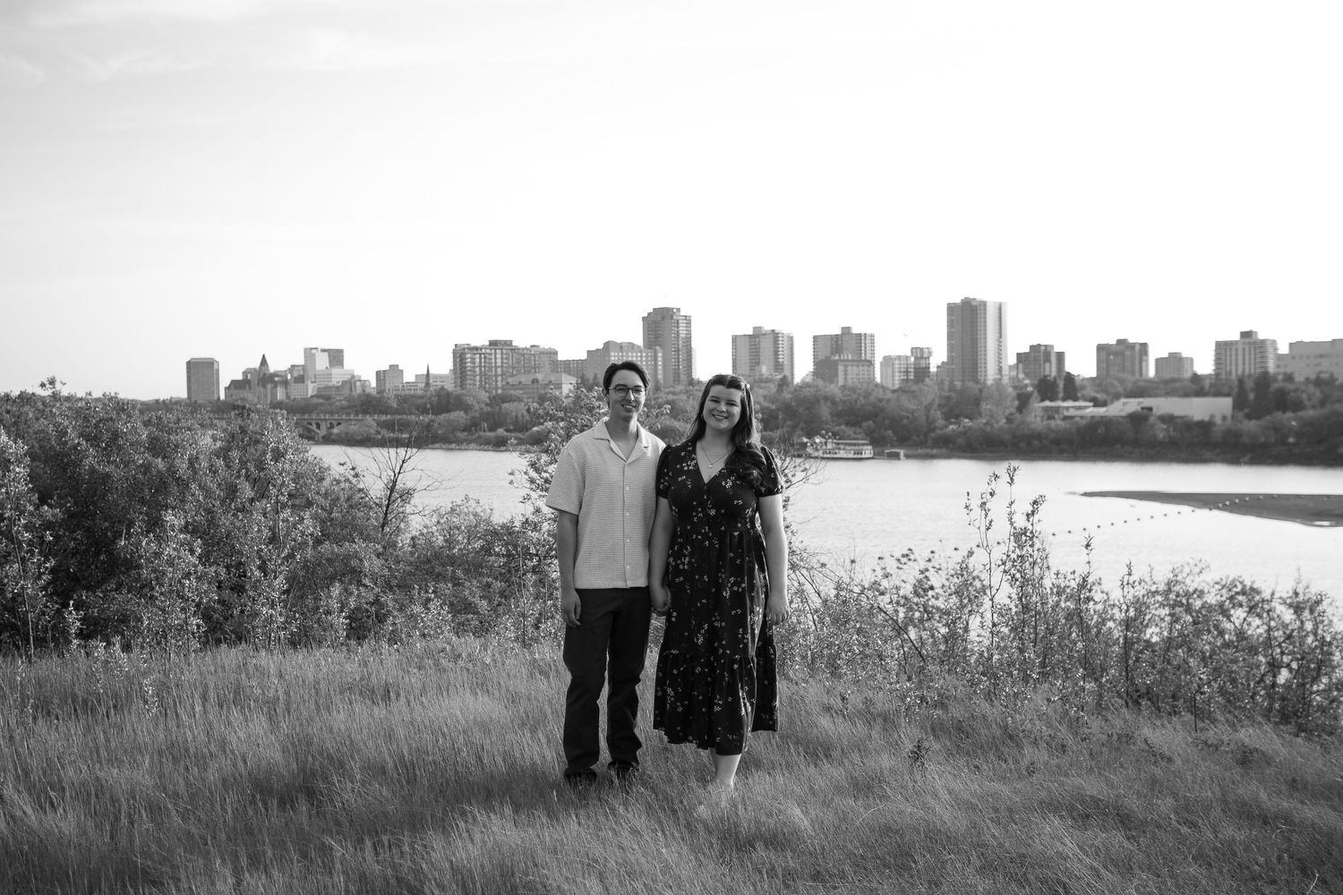 Black and white photo of a couple standing in a field with a city skyline and river in the background.