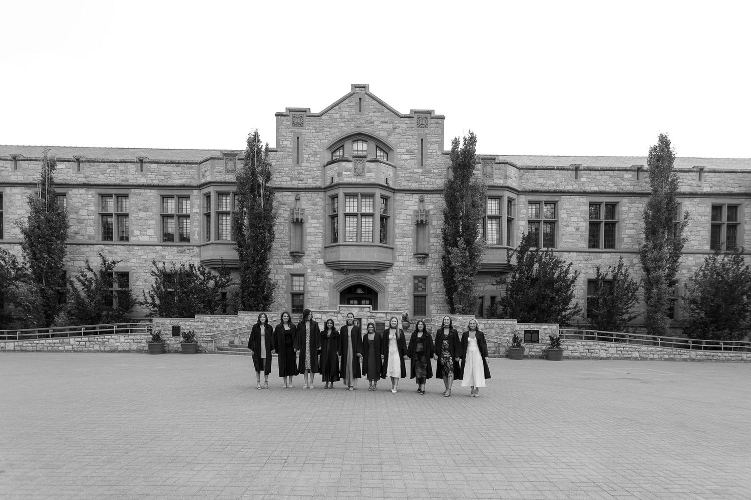 Graduates in caps and gowns pose in front of a historic stone university building in a black and white photo.