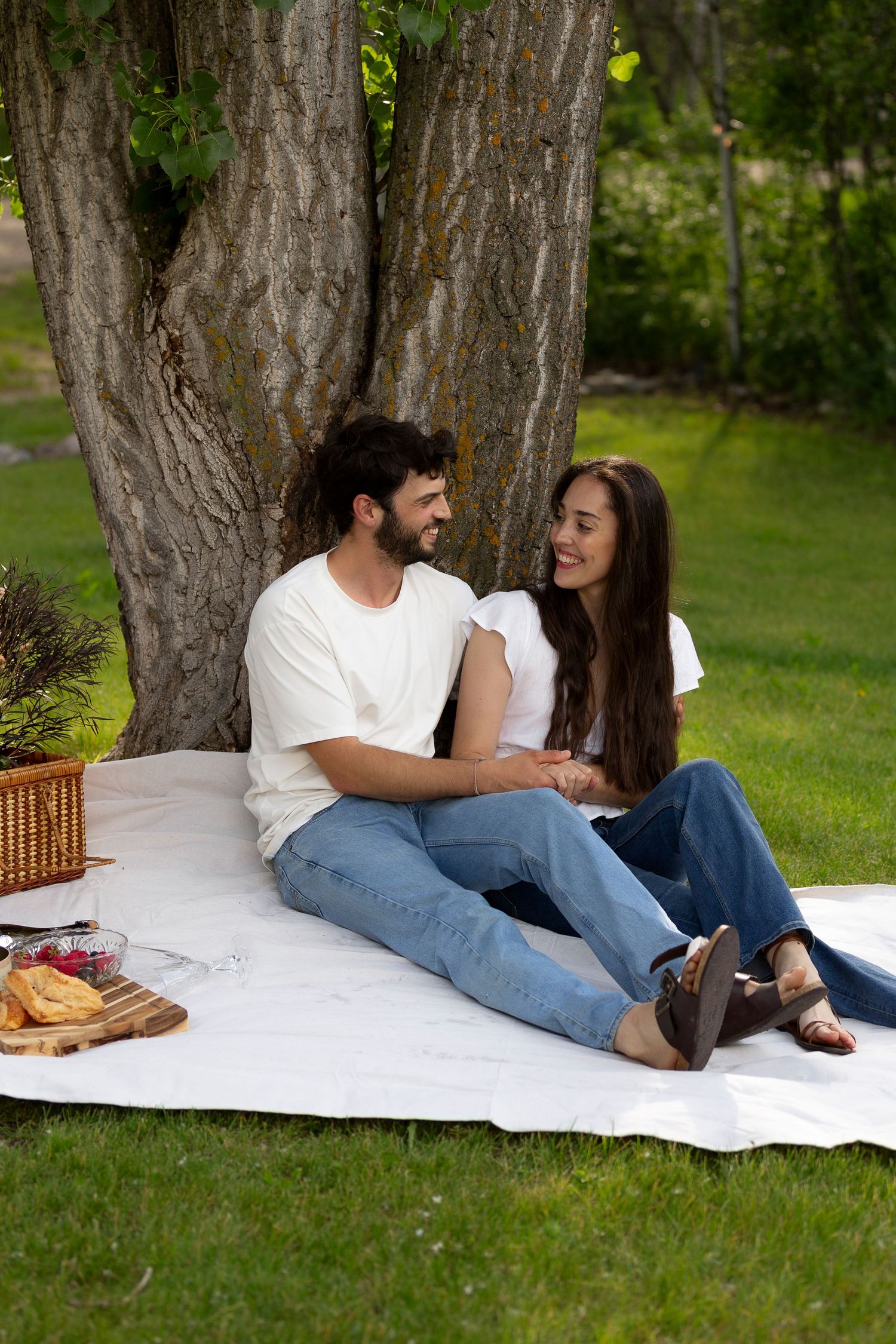Couple sitting on white blanket under large tree enjoying romantic outdoor picnic with basket and food on sunny day.