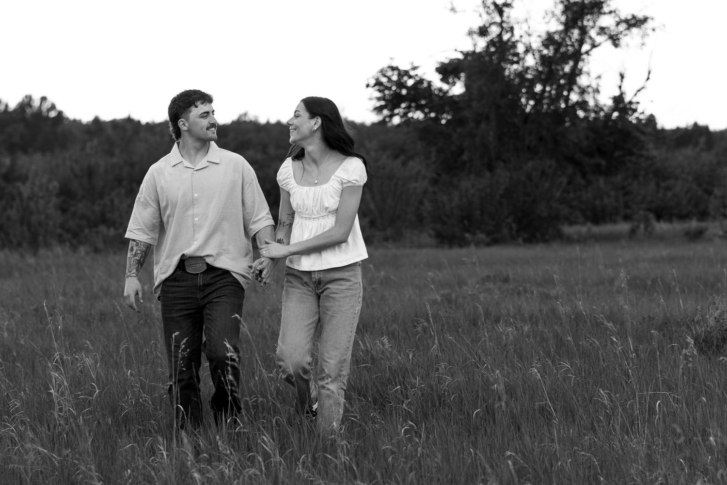 Black and white photo of a couple holding hands and walking through a grassy field with trees in the background.