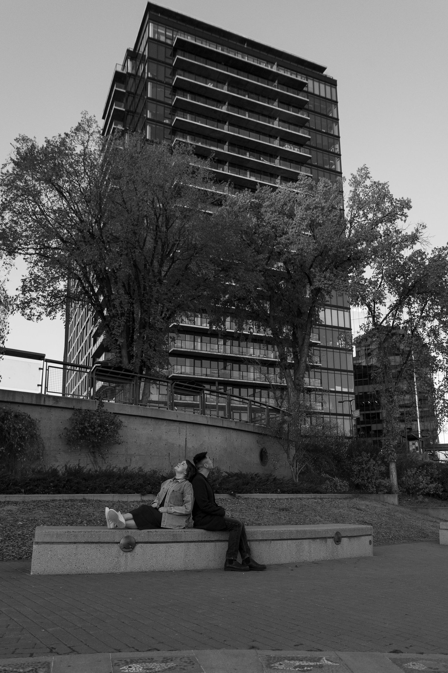 Black and white photo of a couple sitting on a bench in an urban park with a modern high-rise building behind them.