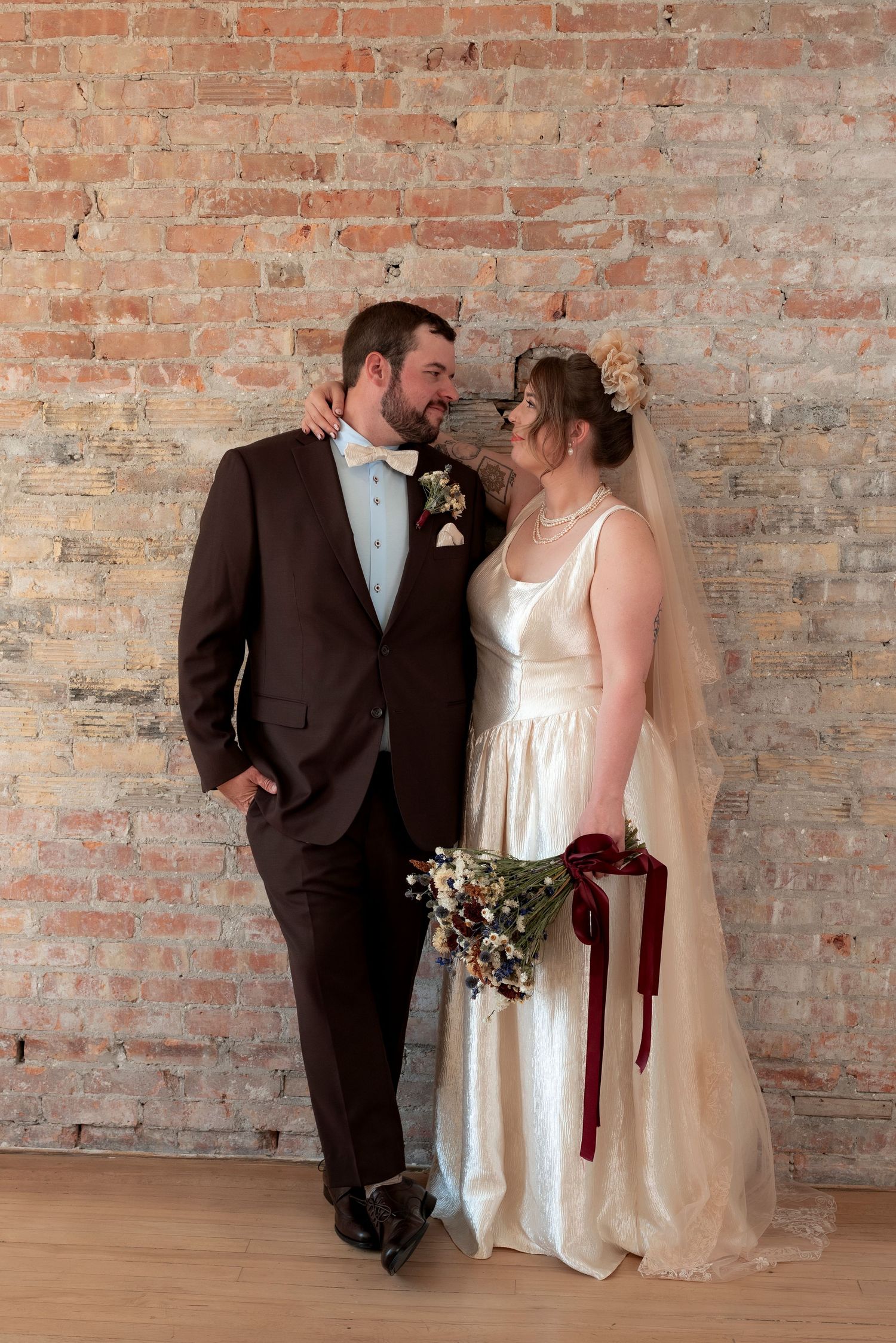 Bride and groom share a romantic moment in front of a rustic brick wall, she holds a burgundy bouquet.