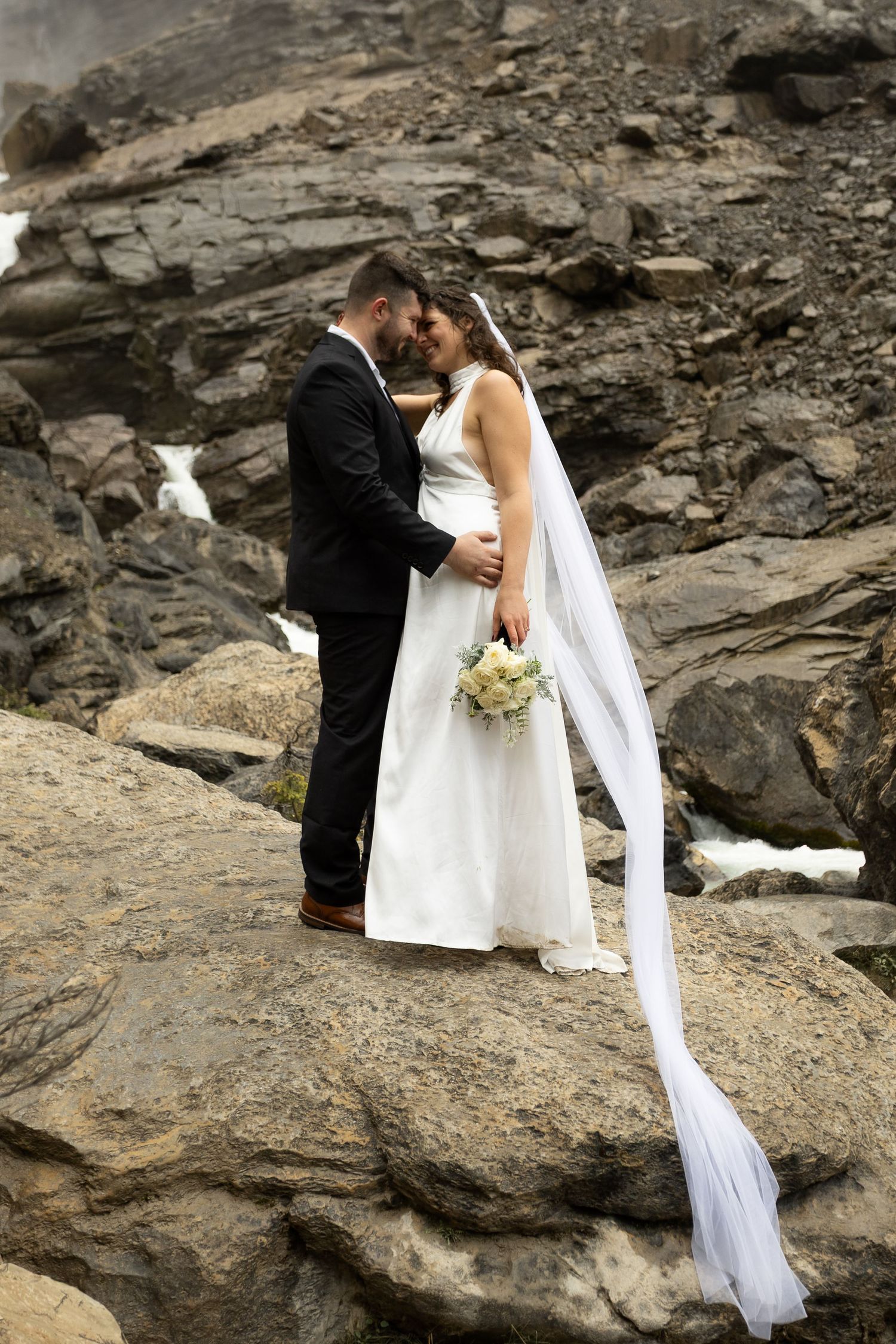 Bride and groom share romantic moment on rocky terrain with waterfall backdrop, bride holding bouquet with flowing veil.