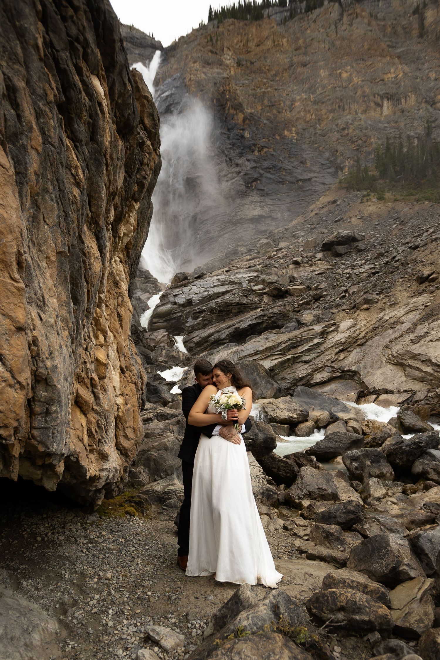 Bride and groom embrace near a stunning waterfall in a rocky mountain canyon, bride holding a white bouquet.