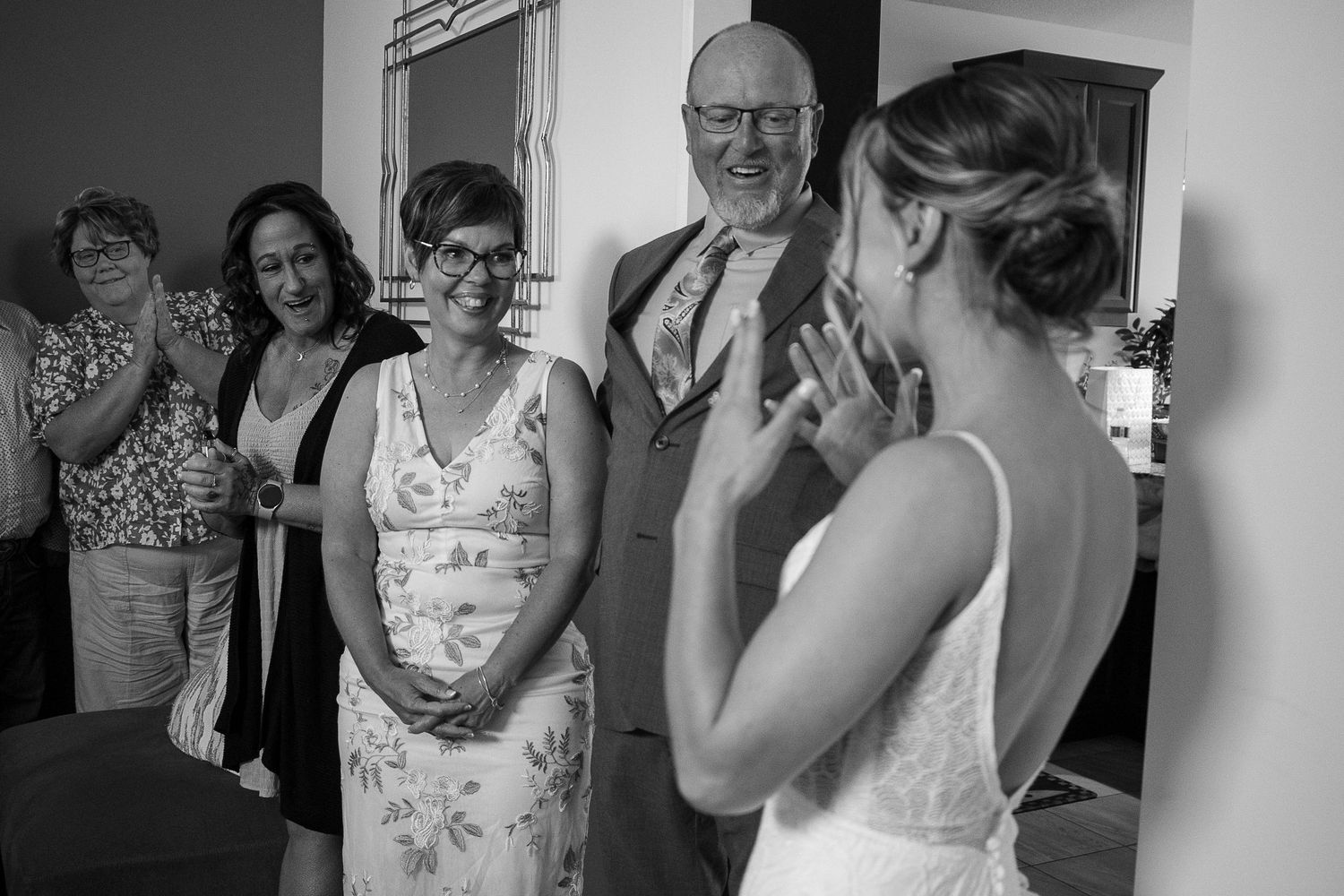 Bride in wedding dress shares emotional first look moment with smiling family members in black and white photo.