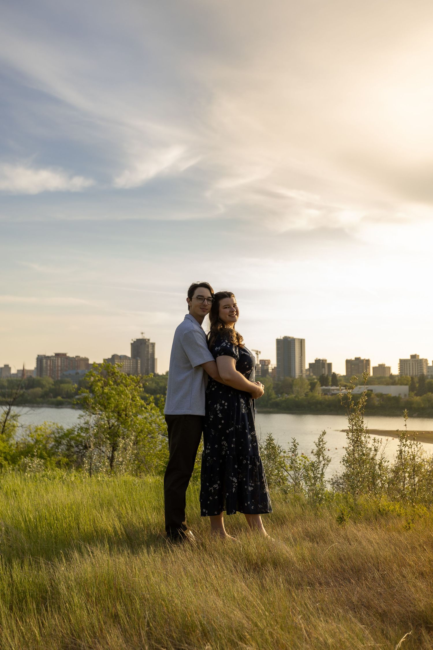 Couple embracing at sunset with city skyline and river in background, standing on grassy hill.
