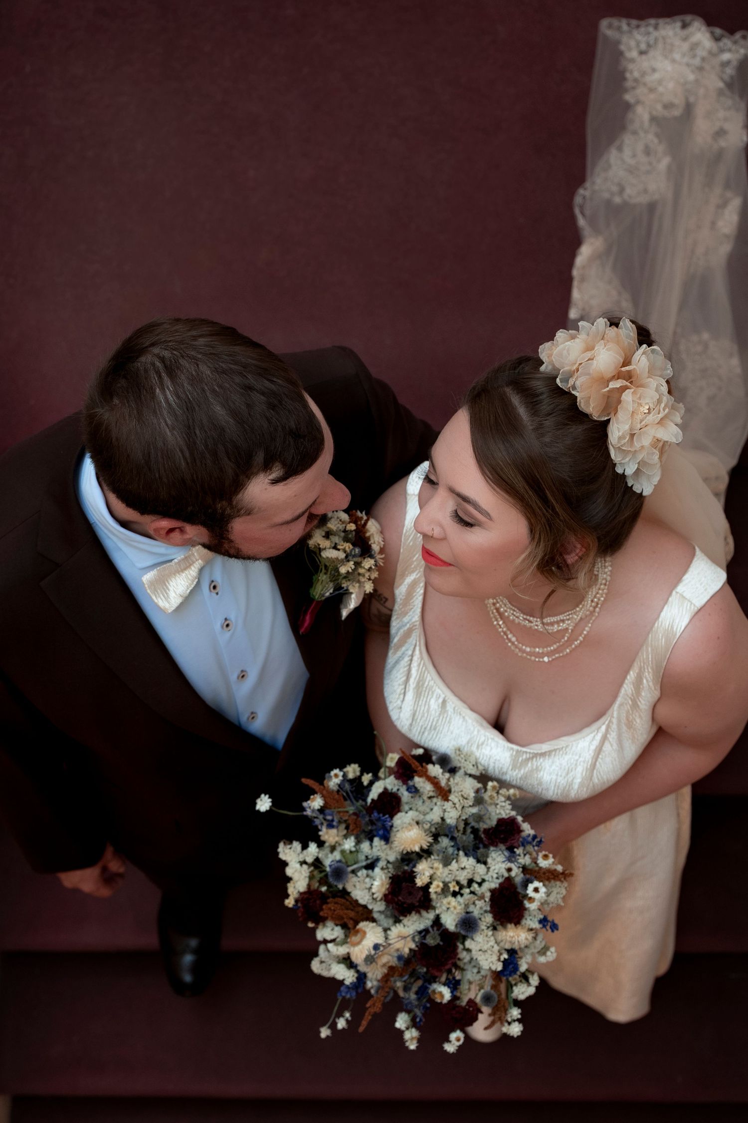 Aerial view of bride holding floral bouquet and groom in bow tie sharing a romantic moment on stairs.