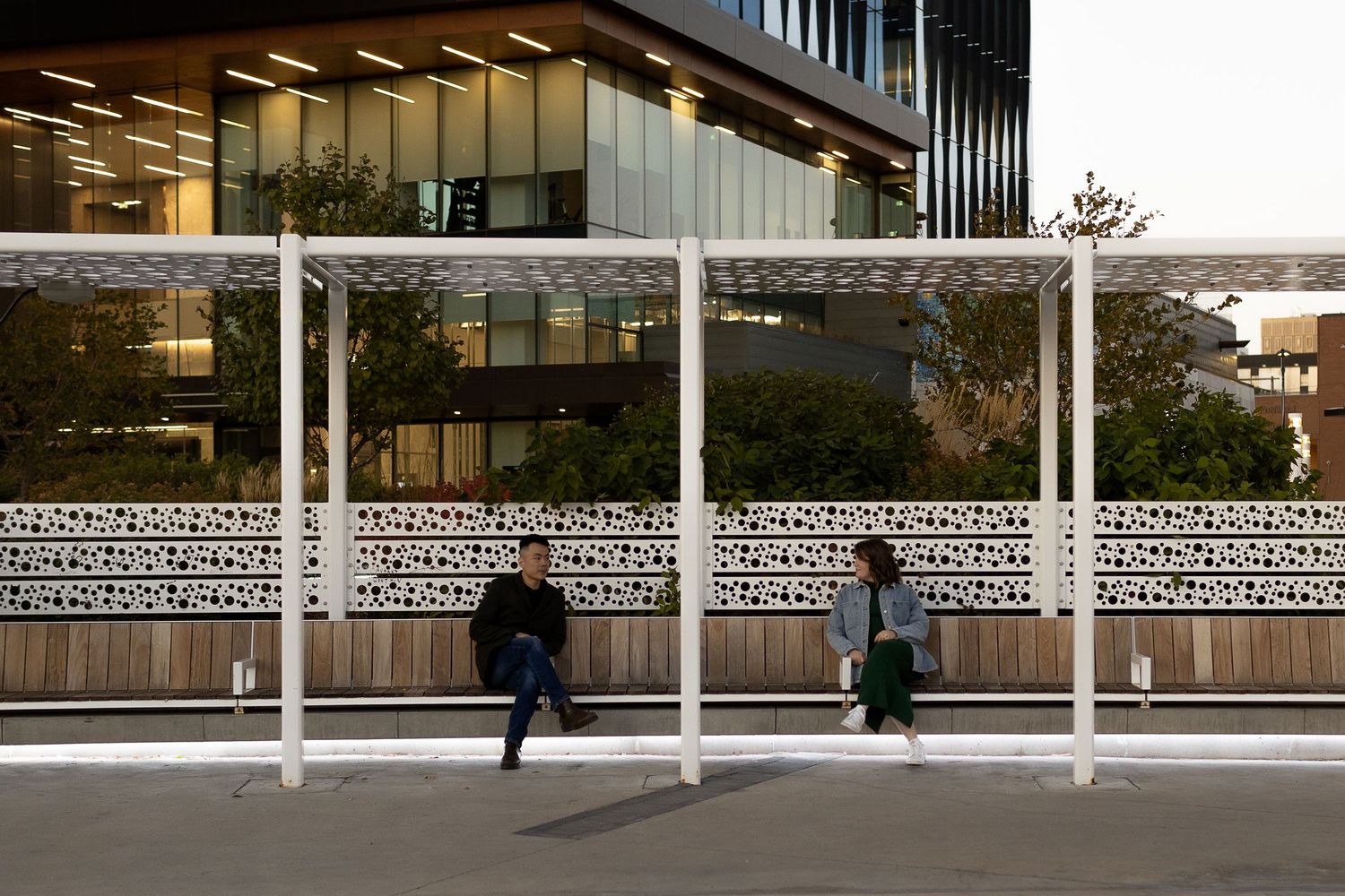 Two people waiting at a modern bus shelter with decorative panels near a contemporary glass office building.