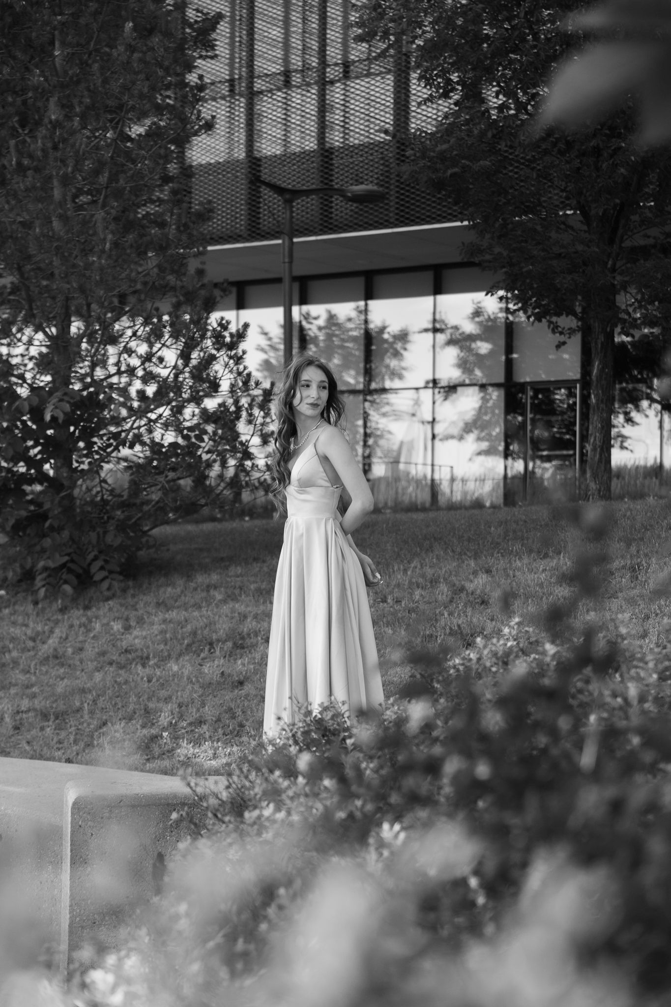 Black and white photo of a woman in a flowing white dress standing in a garden near a modern glass building.
