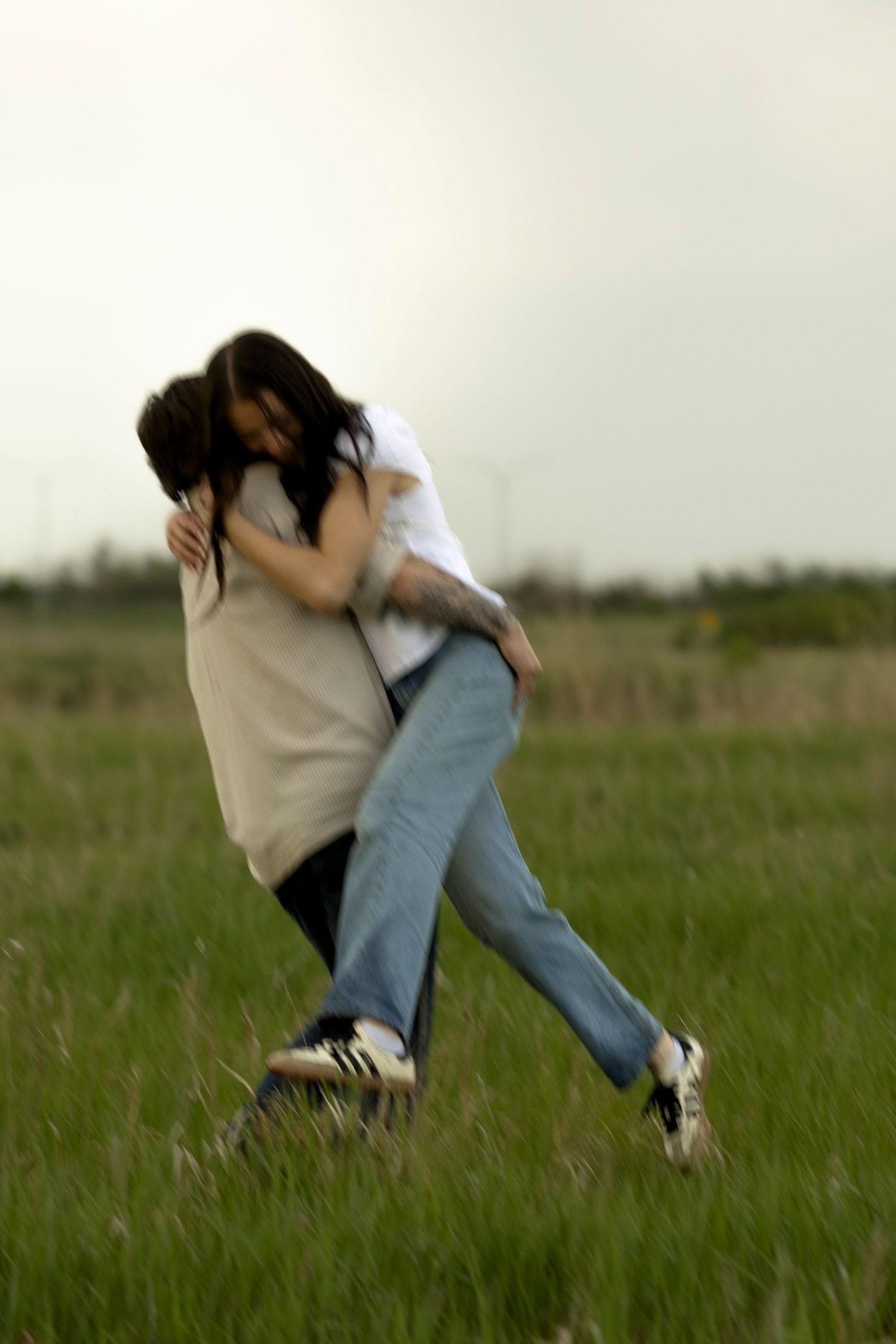 Two people share a romantic embrace in a green field under an overcast sky.