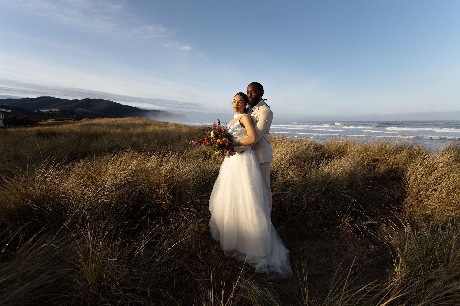 Bride and groom embrace among coastal dunes at golden hour, with ocean waves and rolling hills in the background.