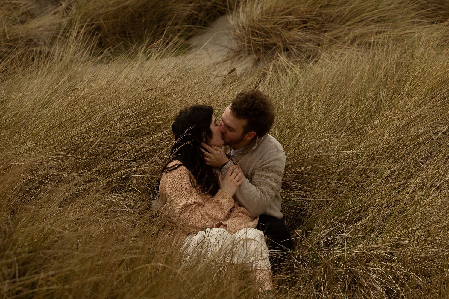 Couple sharing a romantic kiss while sitting together in tall golden grass dunes outdoors.