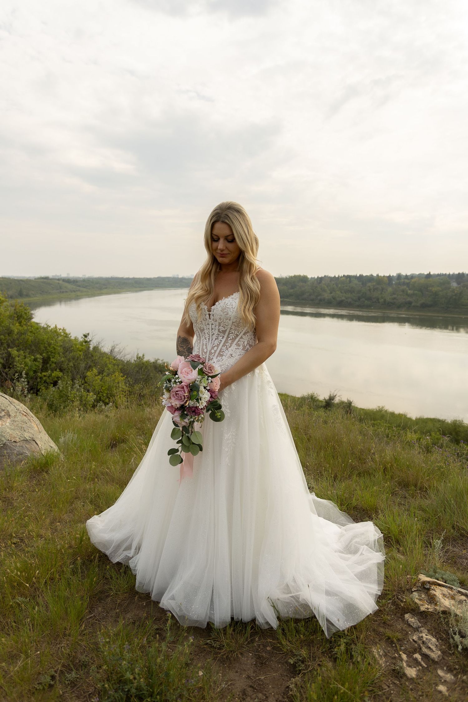 Bride in flowing white gown poses on hillside overlooking serene lake.