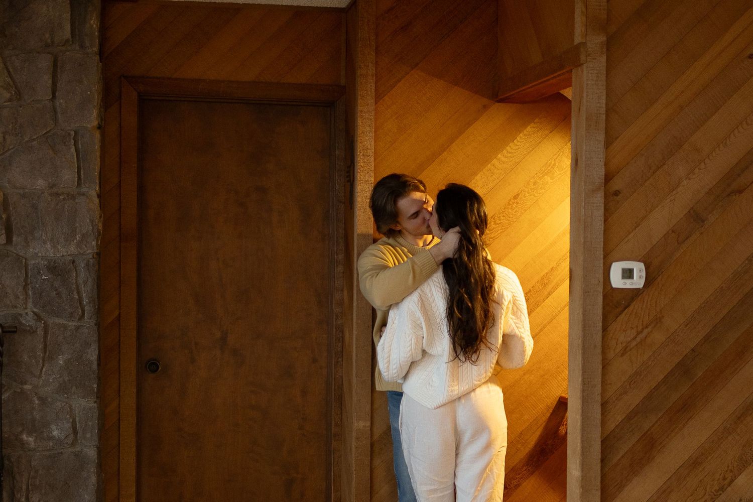 Couple sharing a romantic moment in a warmly lit wooden cabin doorway, dressed in elegant white and yellow outfits.