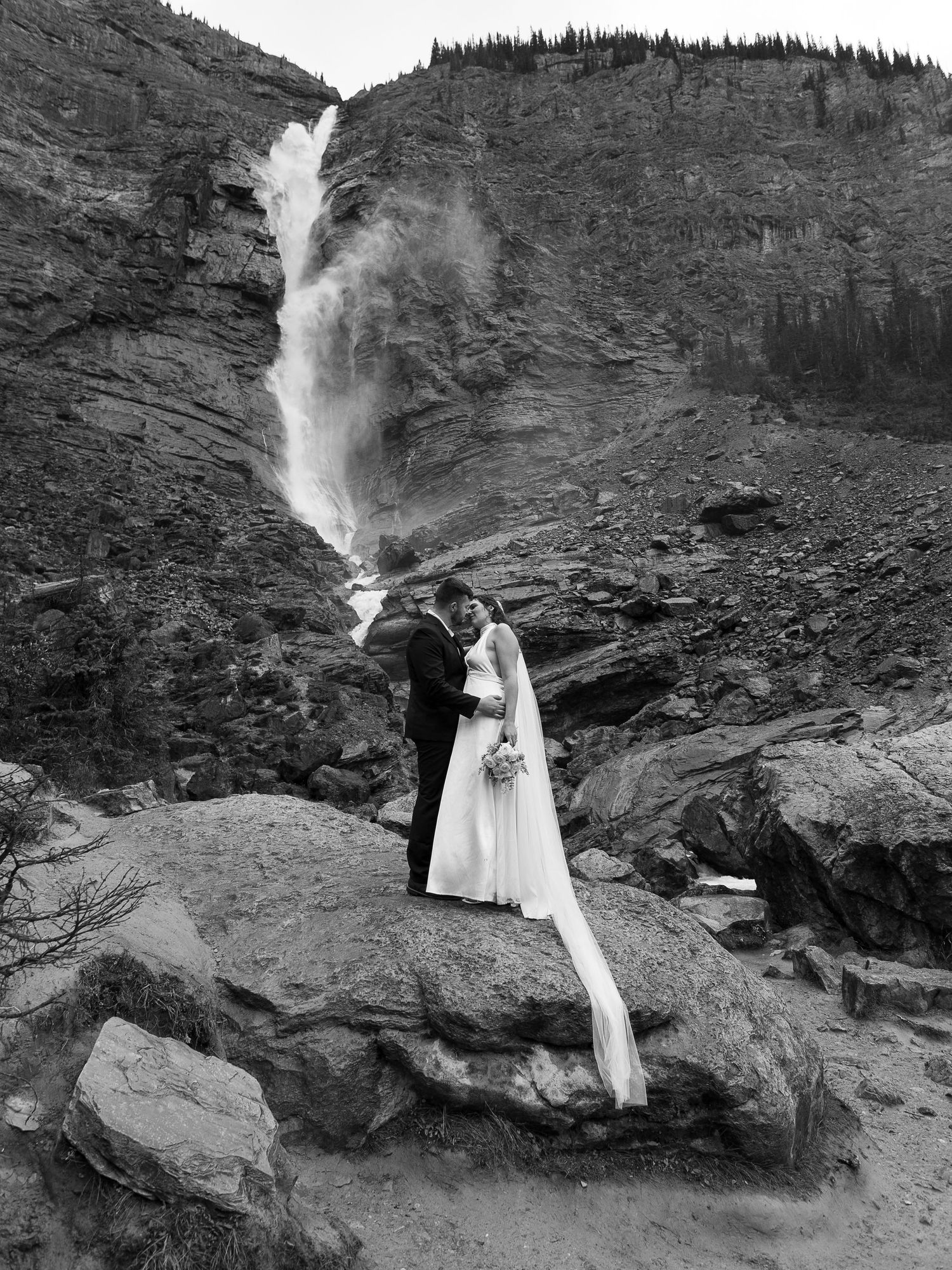 Black and white photo of a bride and groom kissing on rocks before a dramatic waterfall in the mountains.