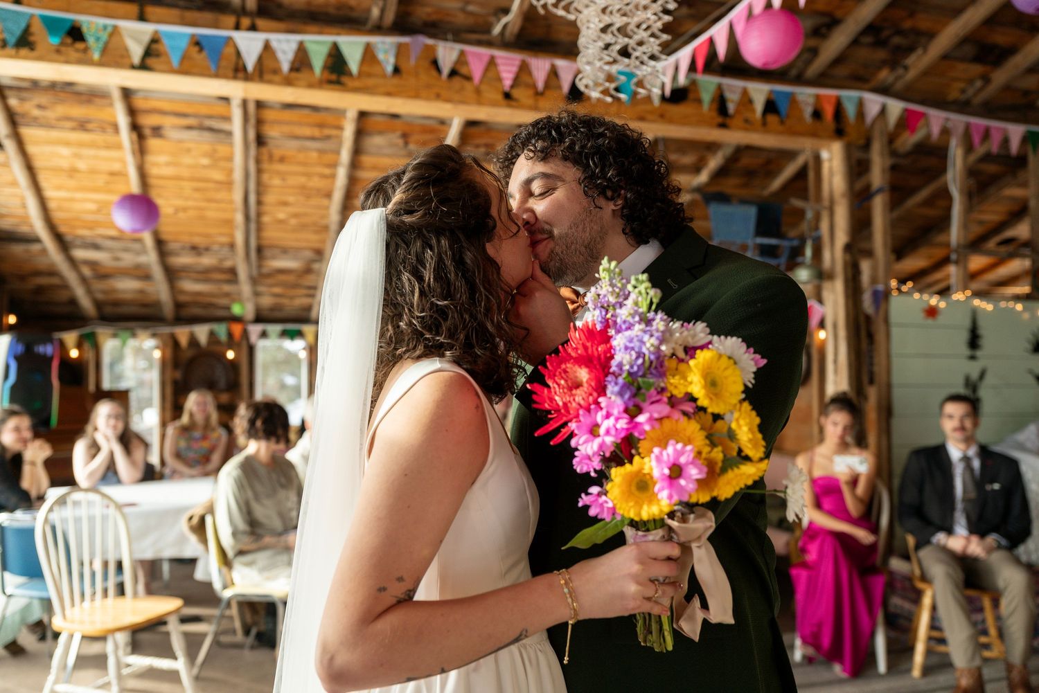 Bride and groom share first kiss at rustic barn wedding, bride holding vibrant bouquet of colorful flowers.