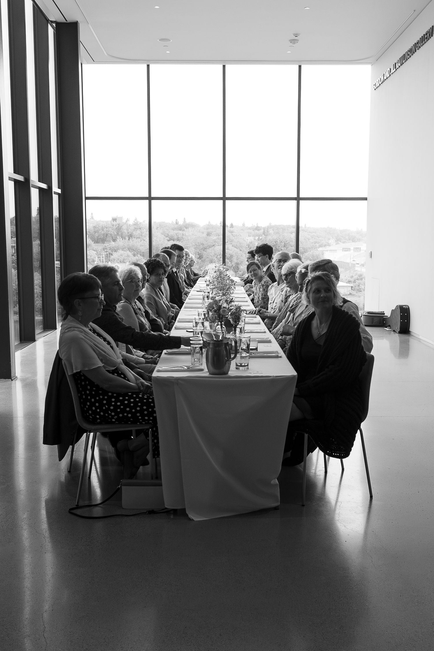 Black and white photo of guests seated at a long dining table near large windows during a formal luncheon event.