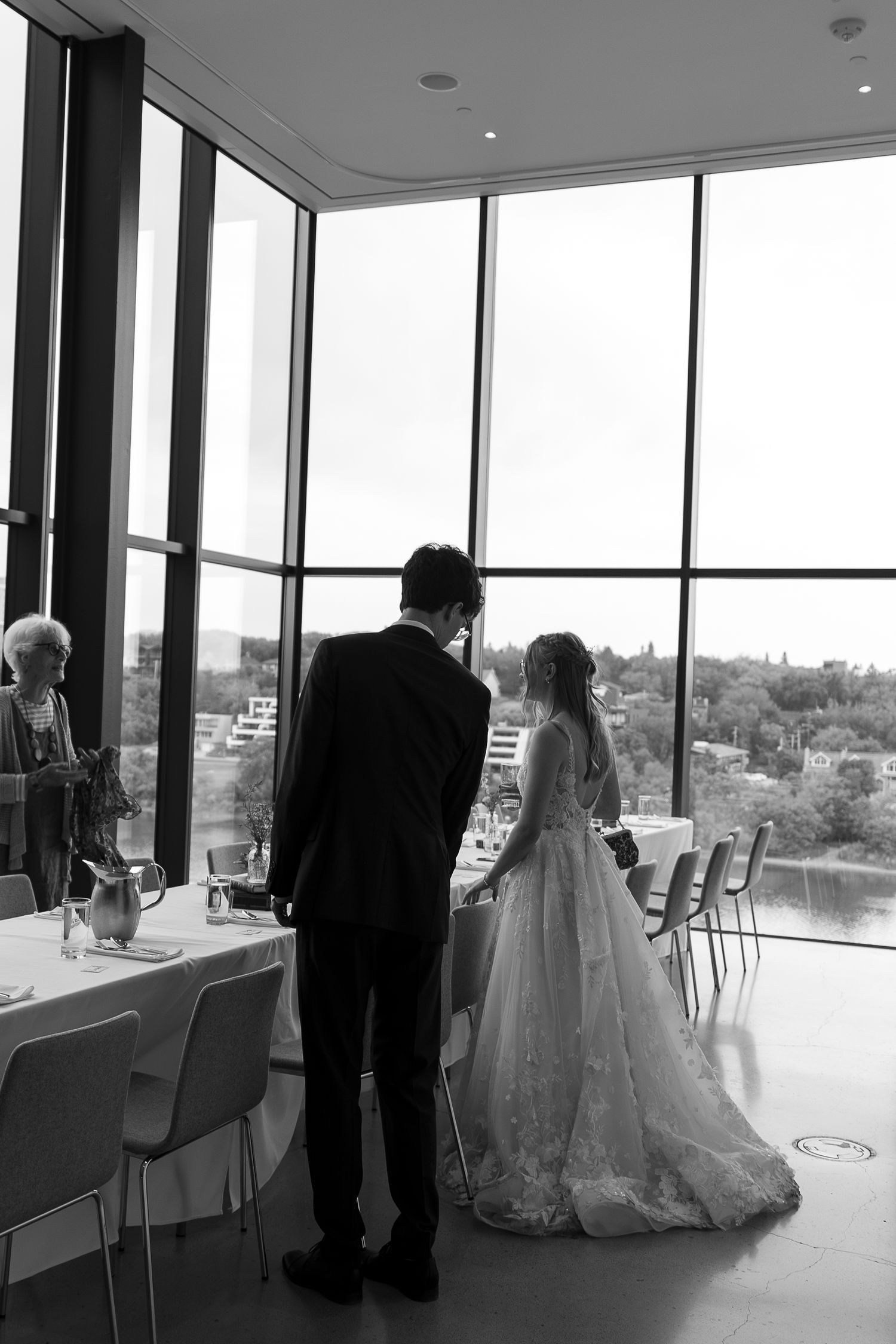 Black and white photo of bride and groom standing together before large windows at elegant wedding venue.