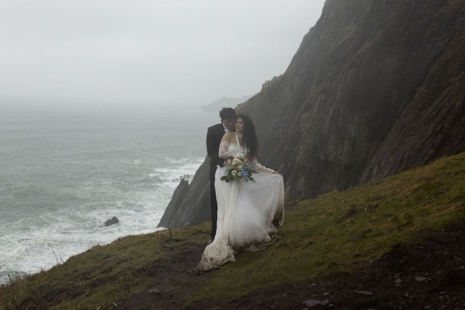 Couple sharing romantic kiss on dramatic misty coastal cliffside, bride holding bouquet in elegant white wedding gown.