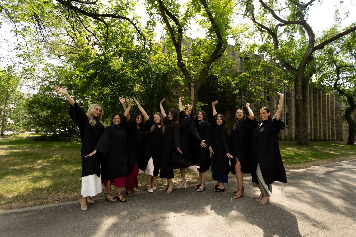 Group of graduates in black robes celebrating outdoors, raising arms joyfully under green trees.