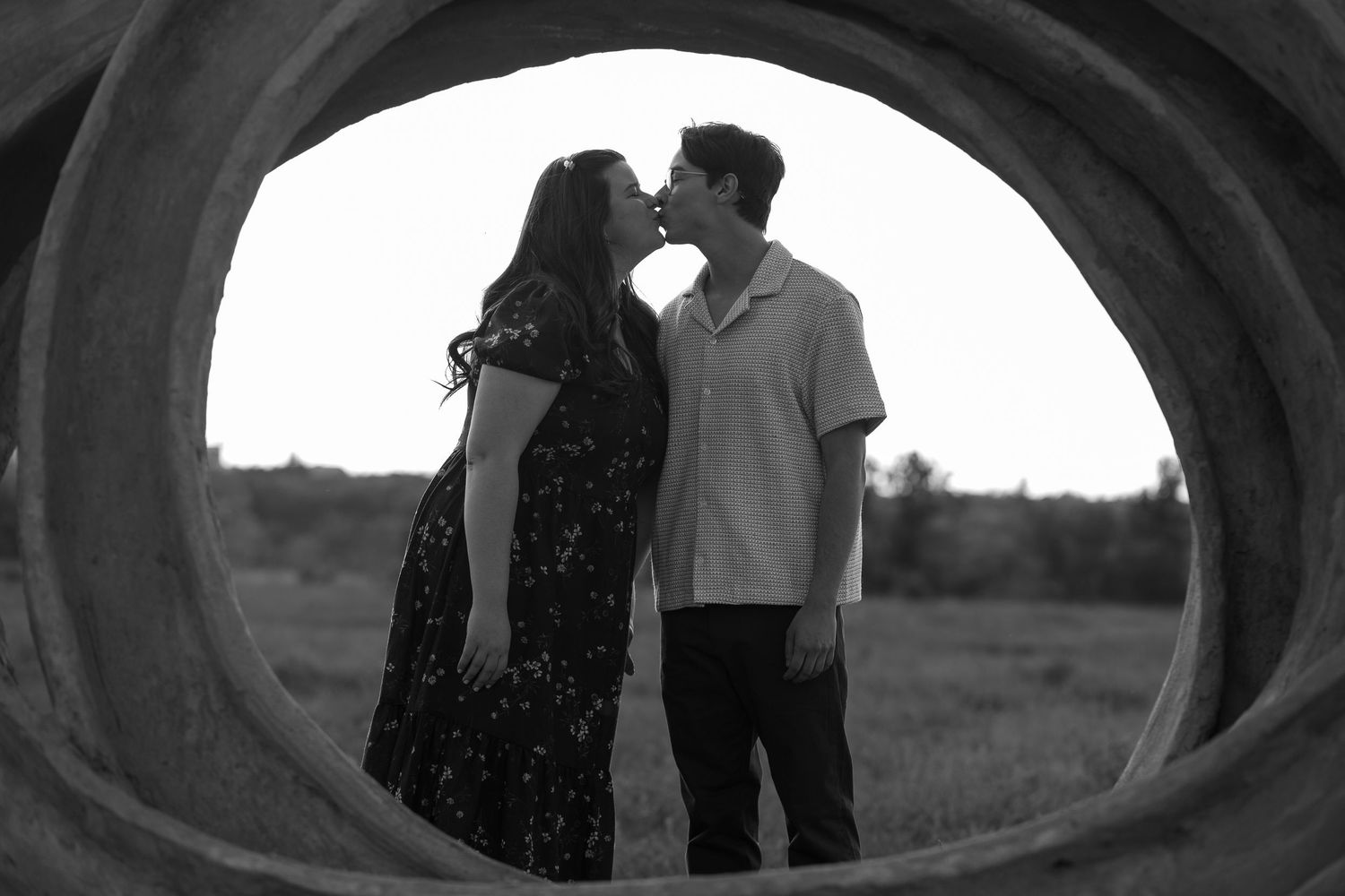 Black and white photo of a couple kissing framed through a circular concrete tunnel in an open field.