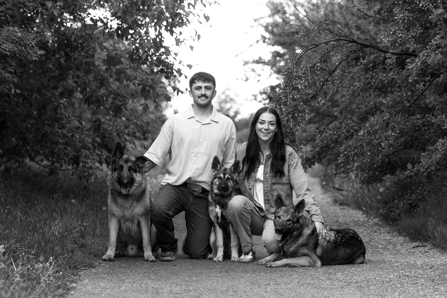 Black and white photo of a couple kneeling on a path with three German Shepherds surrounded by lush trees.